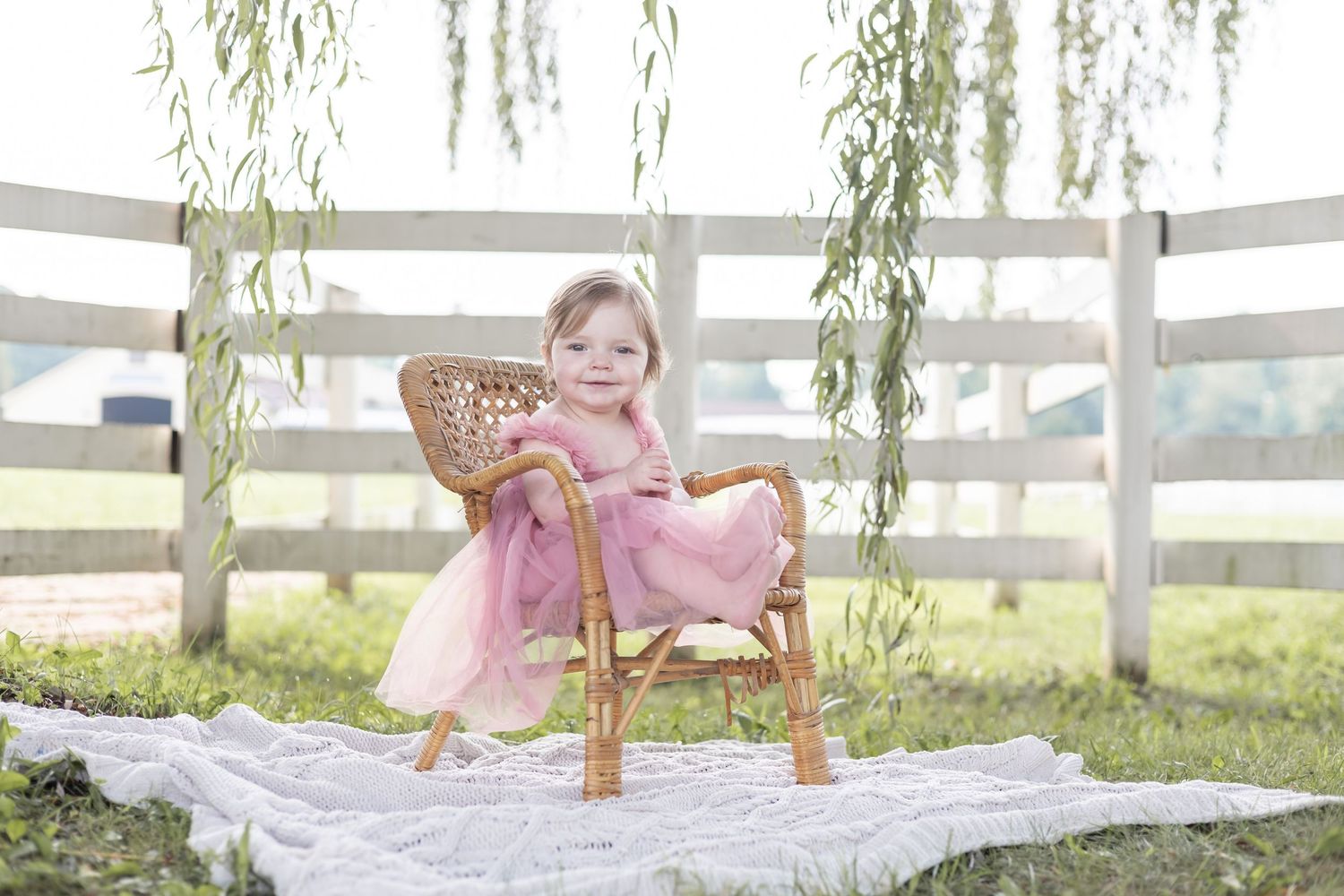 A small figure in a pink dress sits in a wooden chair on a blanket under willow branches by a white fence.