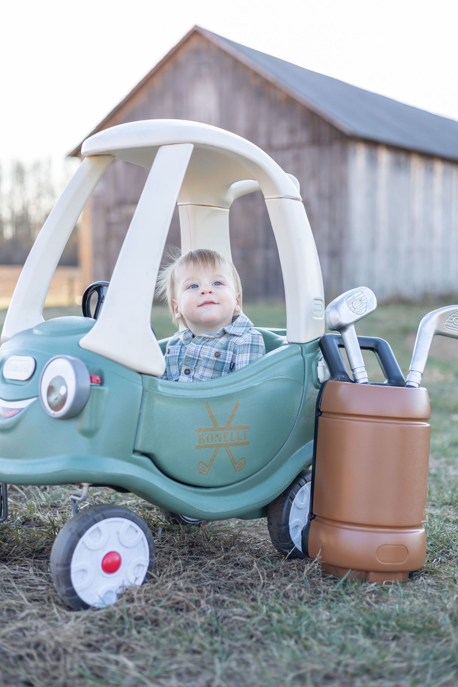 A small green toy cozy coupe car with white roof is parked next to a brown toy golf bag outside a barn.