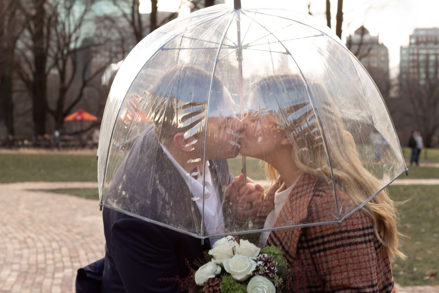 People hold clear bubble umbrellas while walking on a path surrounded by bare trees on a cloudy day.