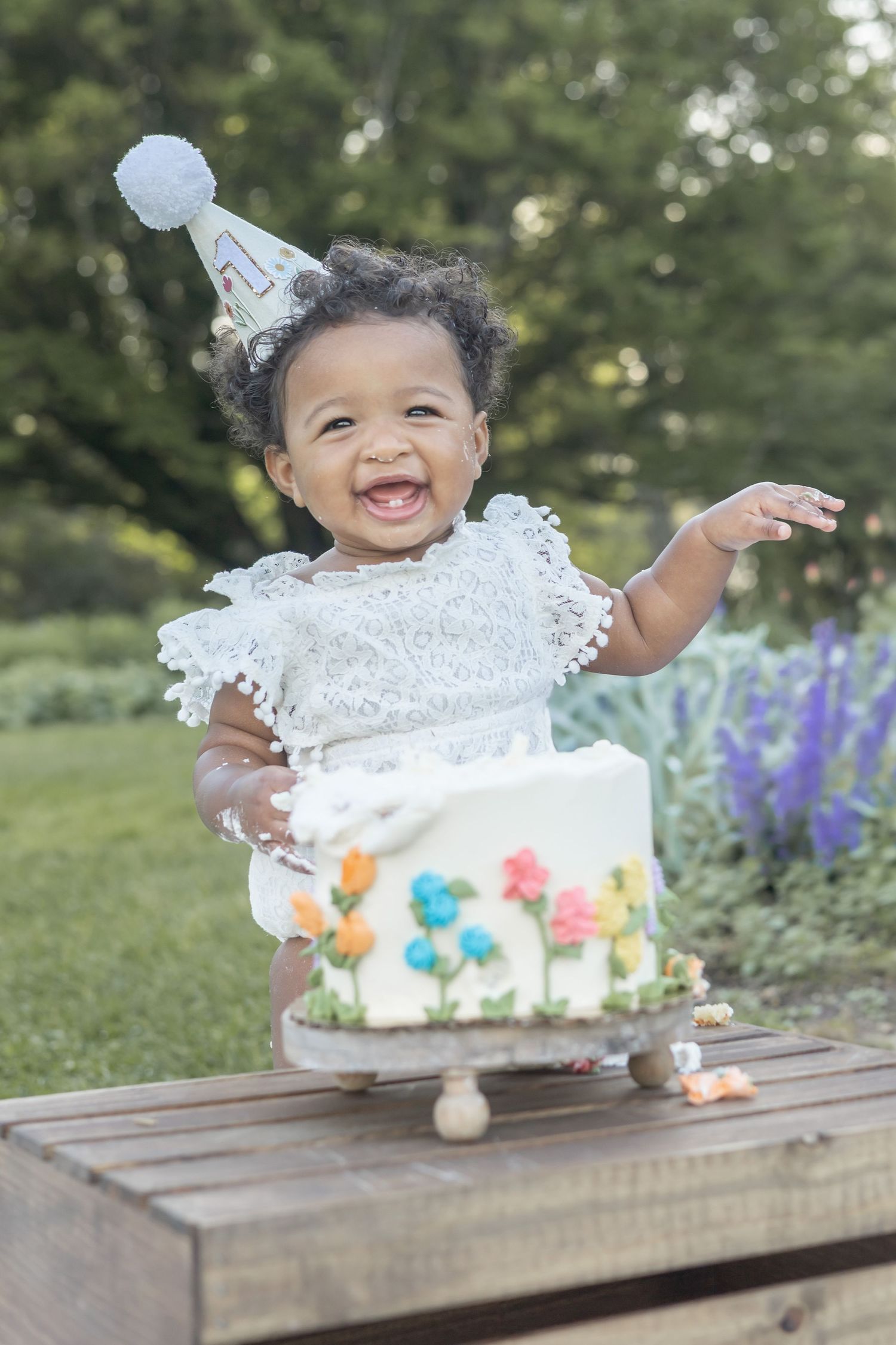 A smiling baby in a white lace dress and party hat celebrates with a decorated birthday cake on a wooden table outdoors.