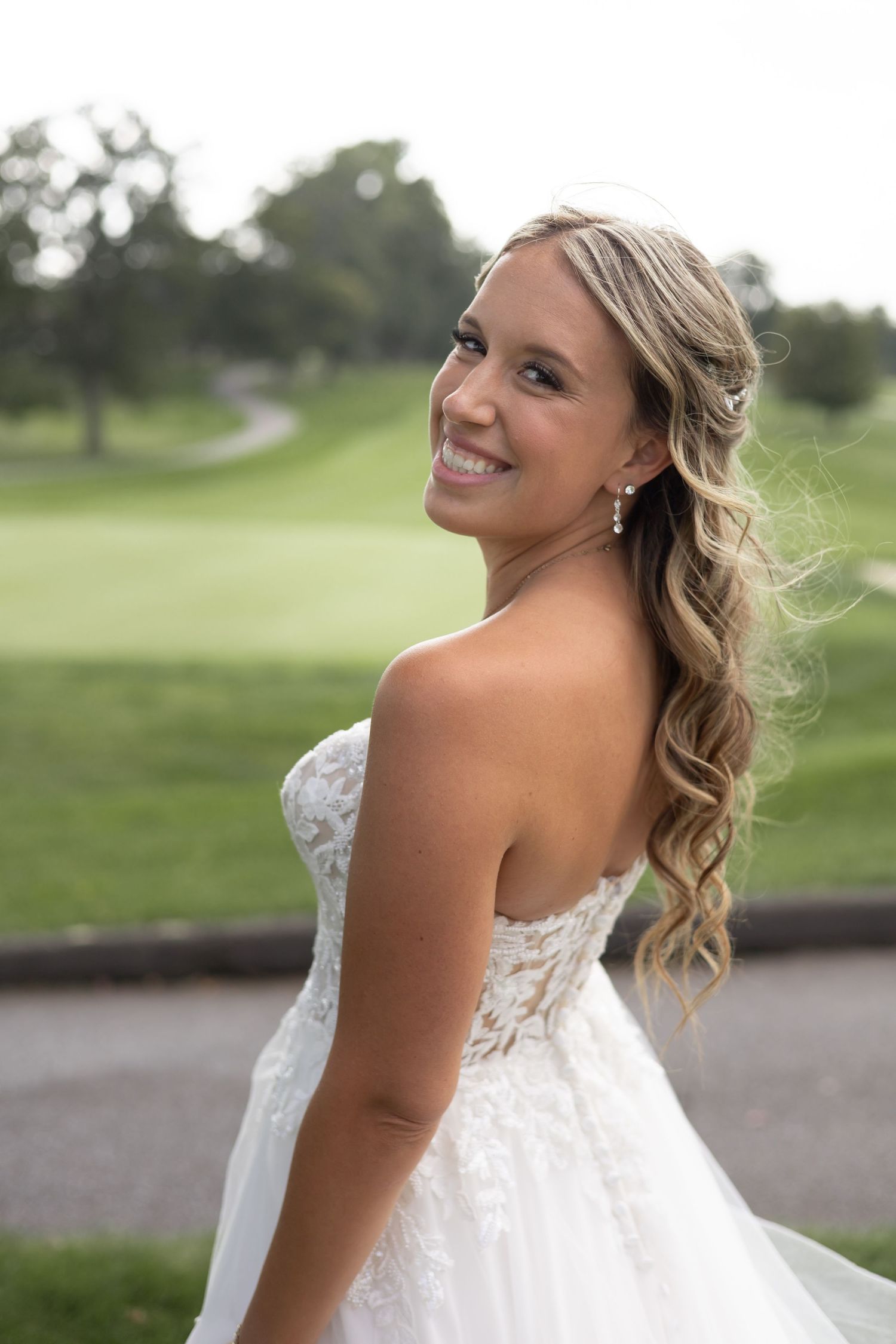 Bride in elegant lace wedding dress poses outdoors on a golf course with curled blonde hair styled to the side.