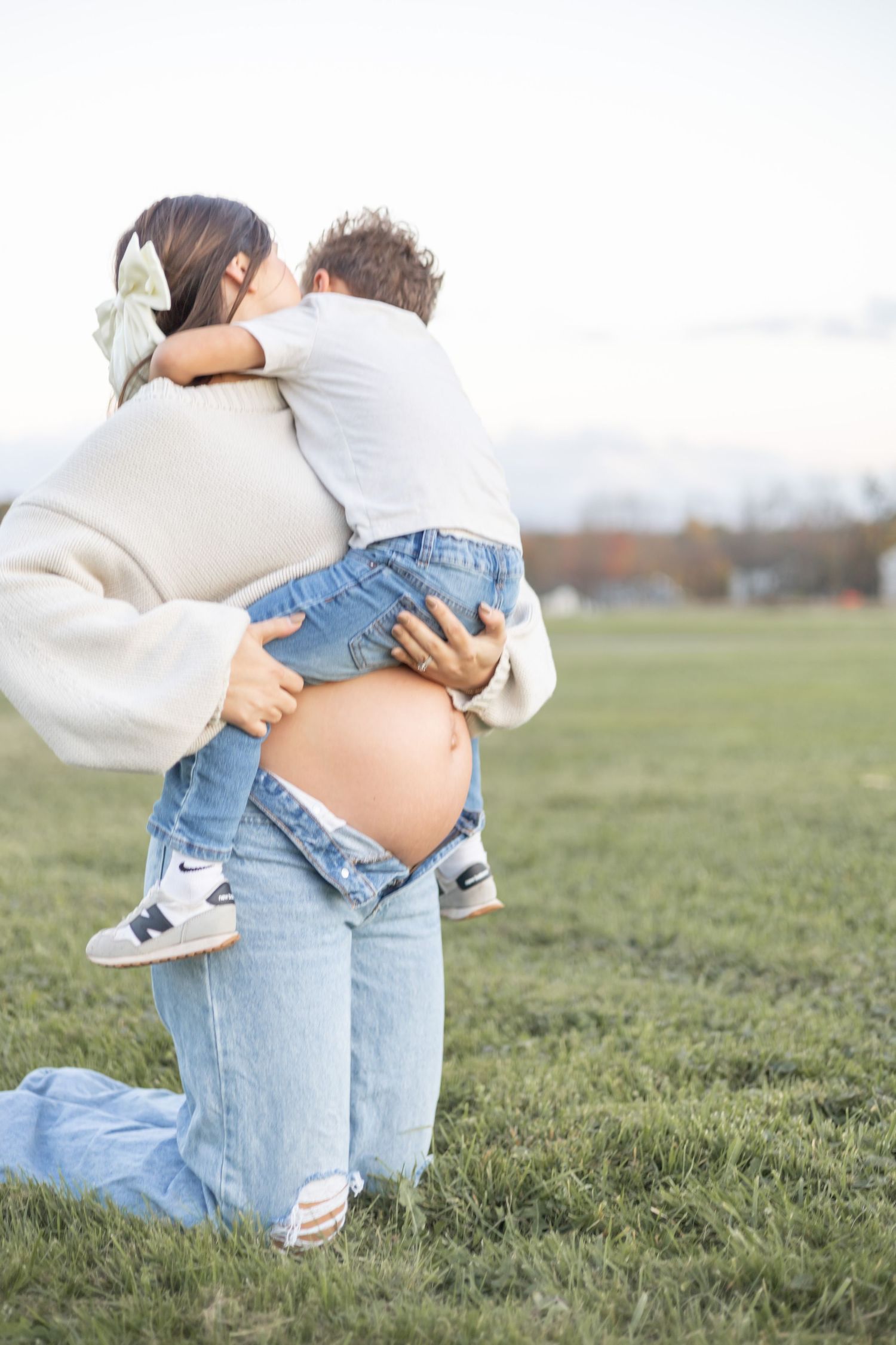 Side view of pregnant woman in sweater holding toddler outdoors in grassy field.