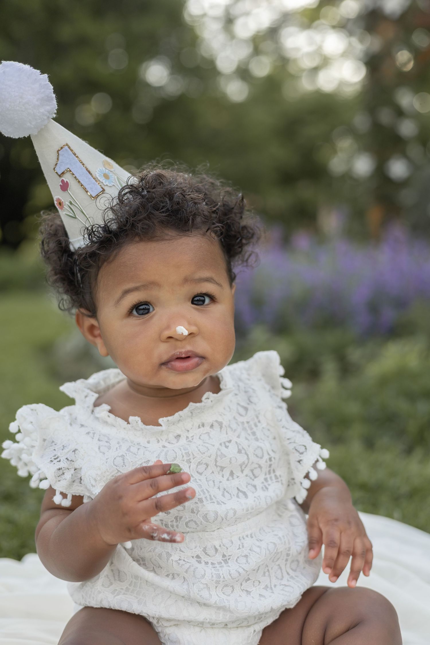 Baby in white outfit and party hat plays while sitting outside with purple flowers in background.