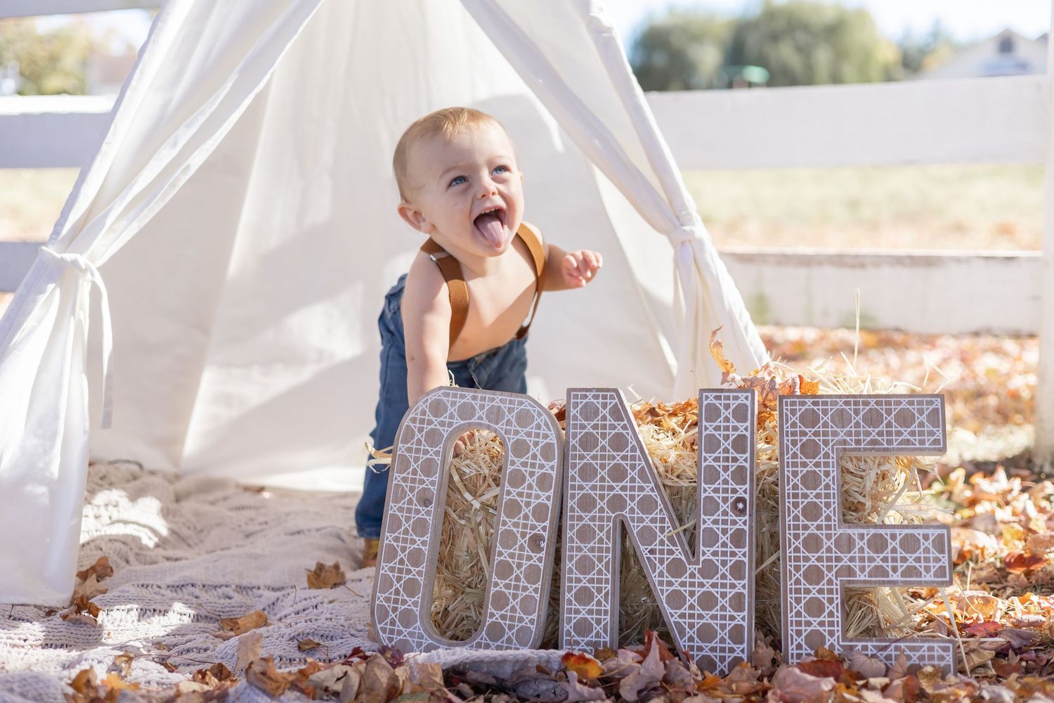 Laughing baby sits behind decorative ONE letters in front of white teepee tent outdoors with autumn leaves scattered around.