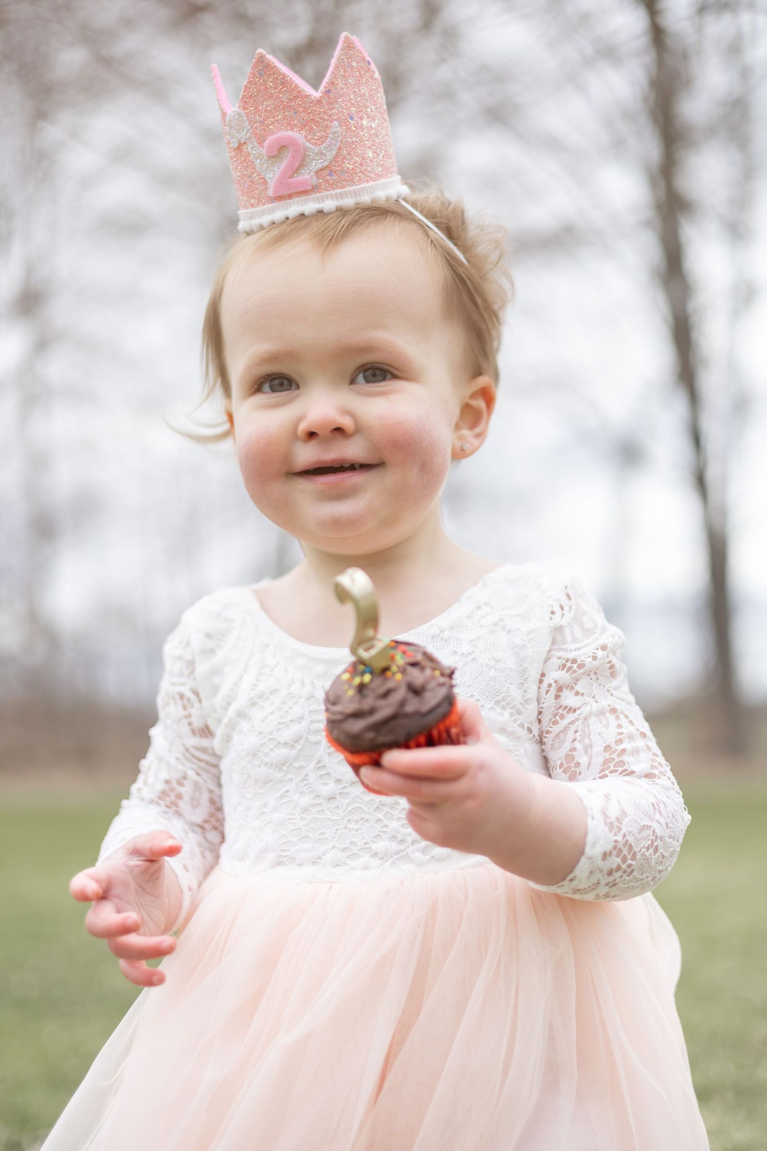 A small figure wearing a pink crown and lace dress holds a chocolate cupcake while celebrating their second birthday.