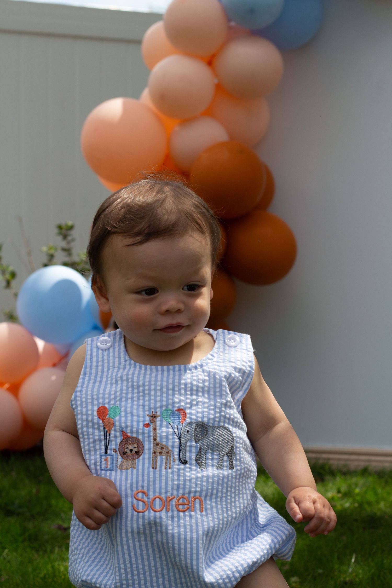 Baby in blue seersucker outfit with balloon decorations in orange and blue colors in the background.