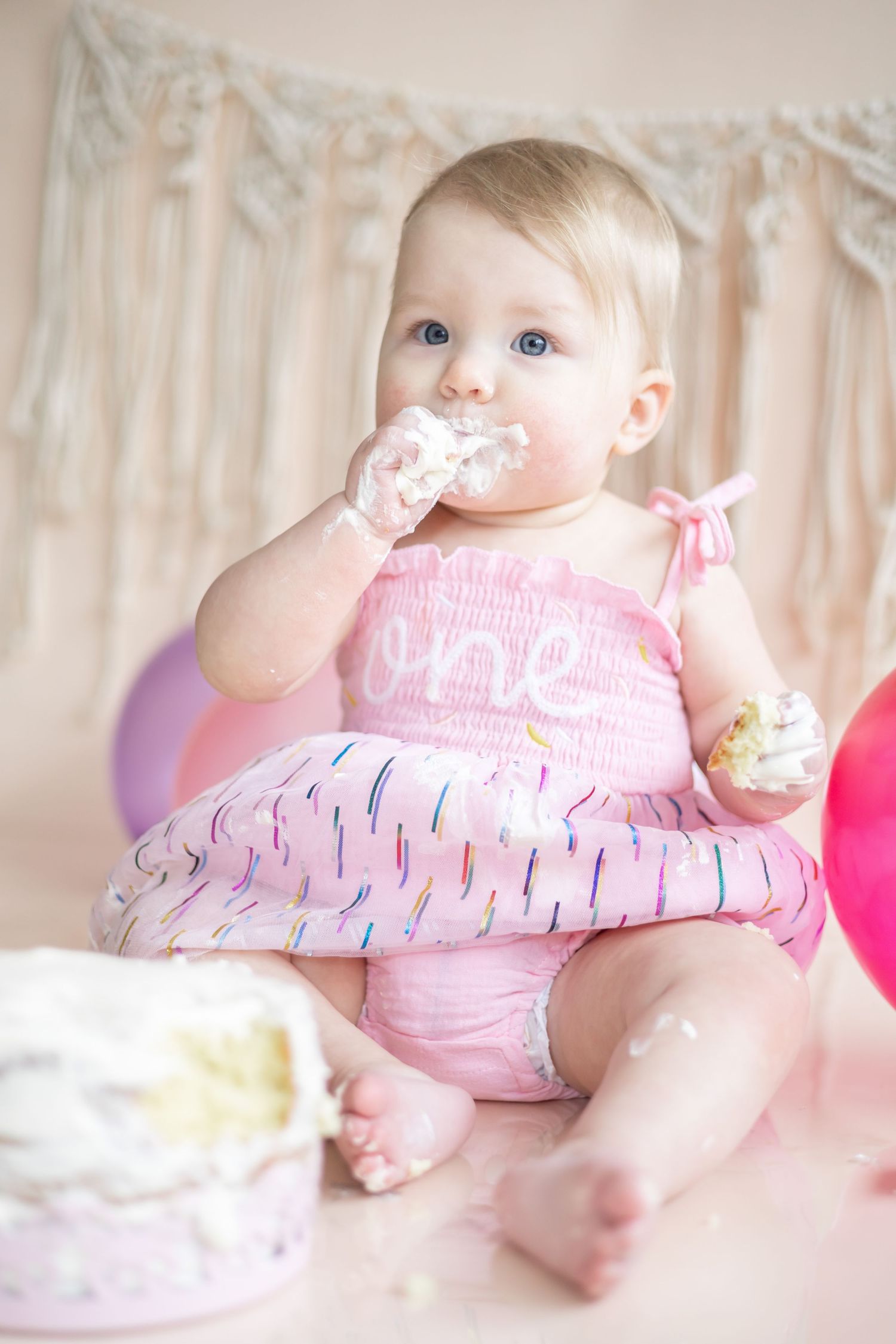 Baby in pink dress smiles while playing with favorite toy in dreamy pastel-colored nursery setting.
