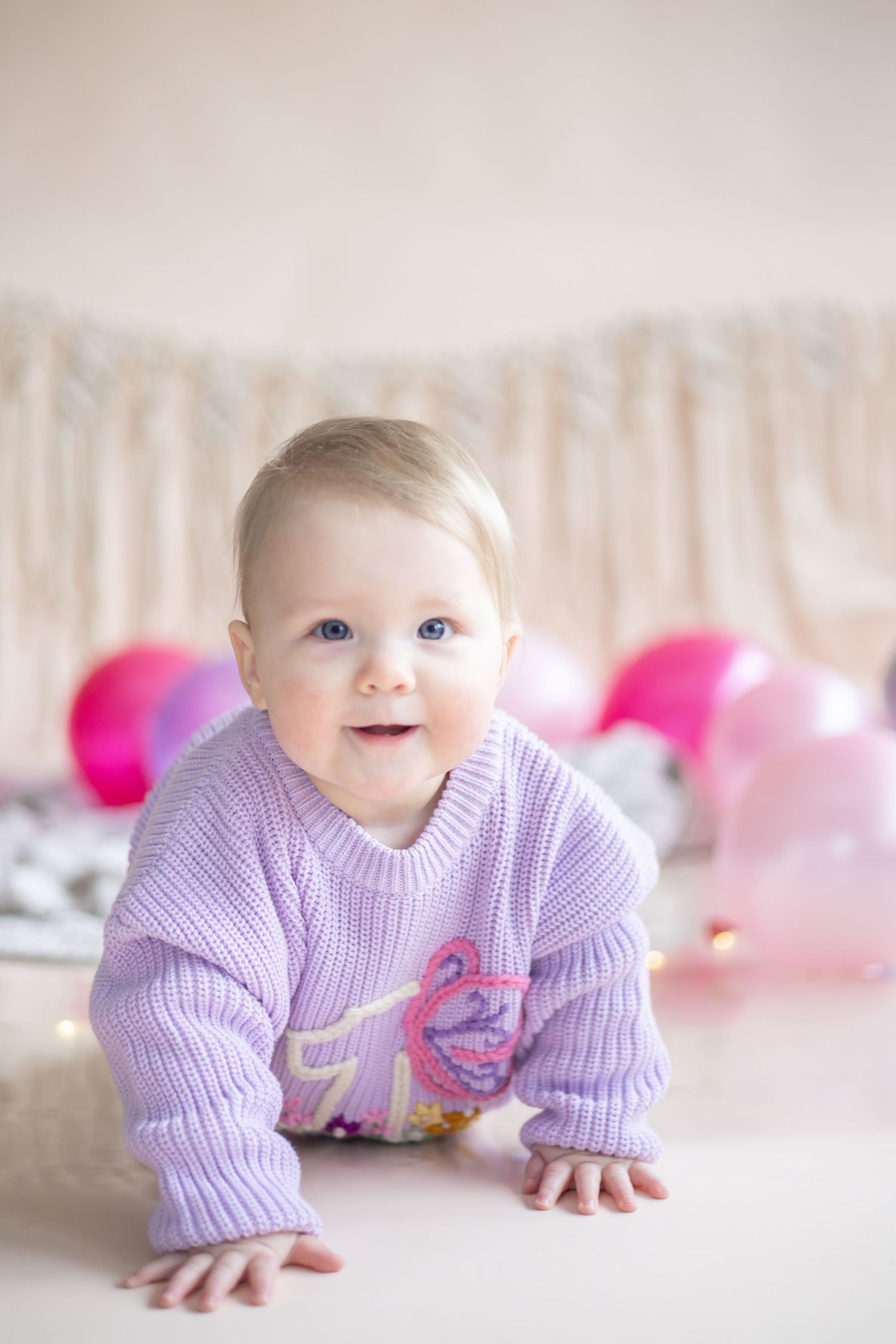 Baby in purple sweater crawls on floor with pink balloons in background during photo session.
