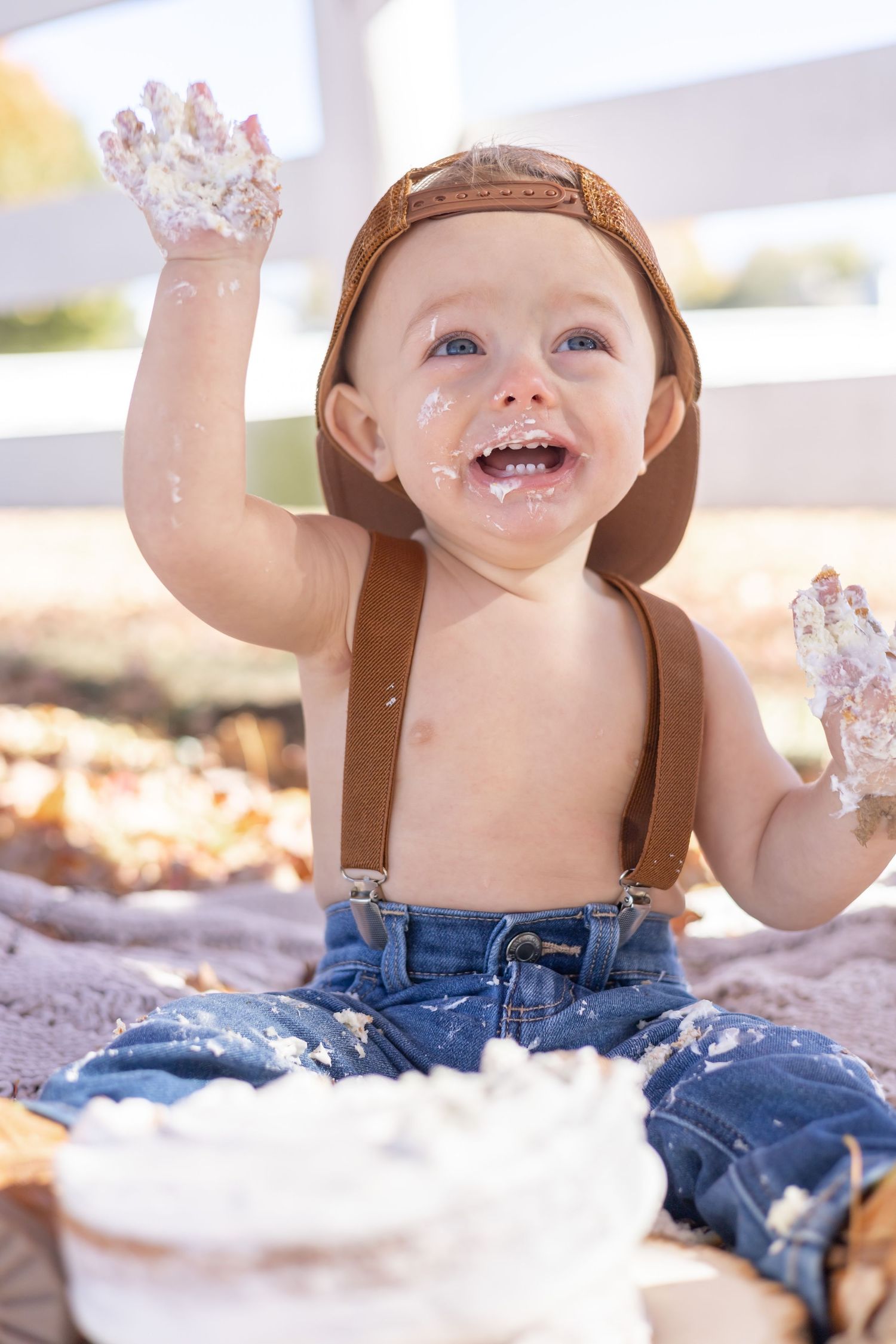 Baby wearing denim overalls and brown hat plays joyfully in white fluffy material with a bright smile and raised hands.