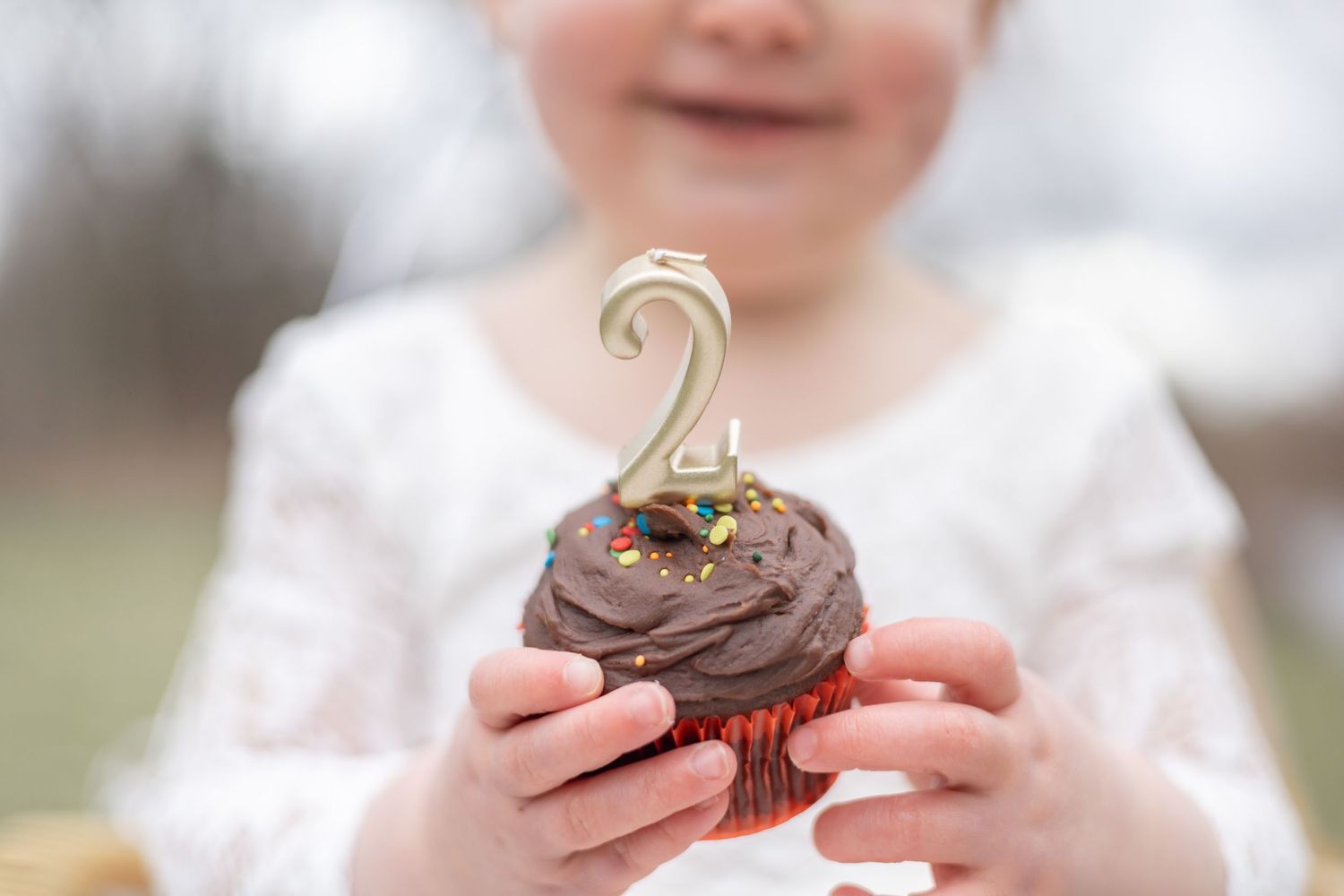 Person smiling while holding a chocolate frosted cupcake with gold number topper decoration.
