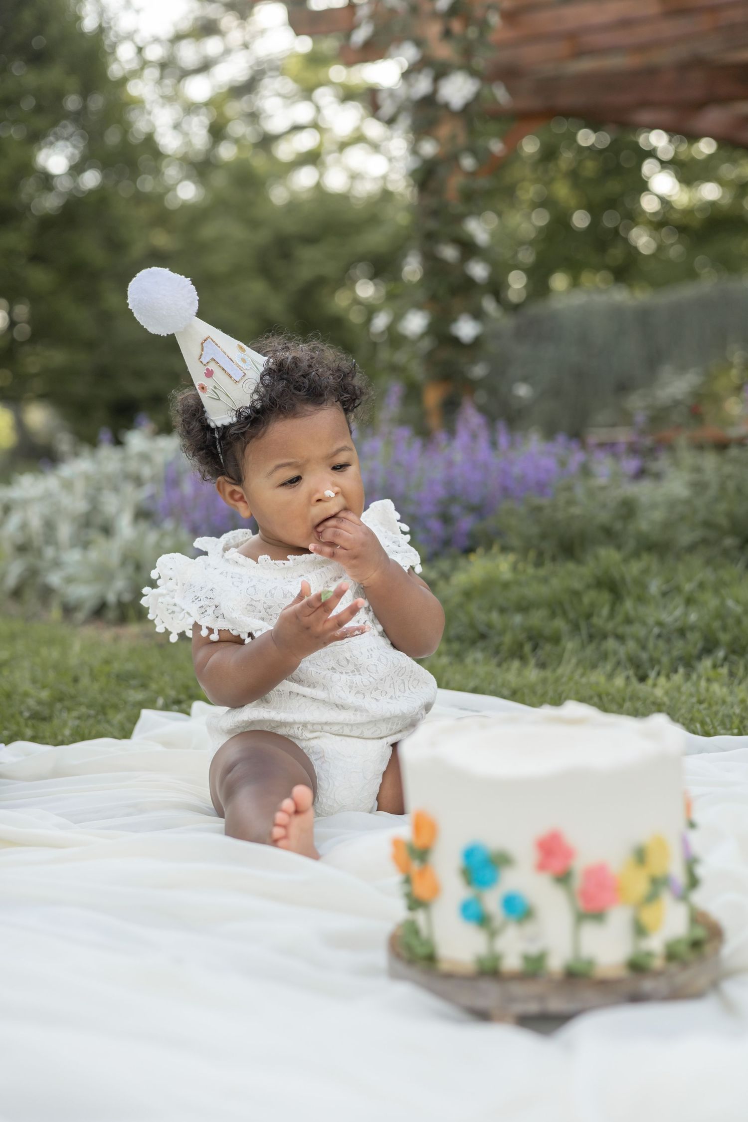 Baby in white dress smashing face into birthday cake during outdoor cake smash photo session with purple flowers.