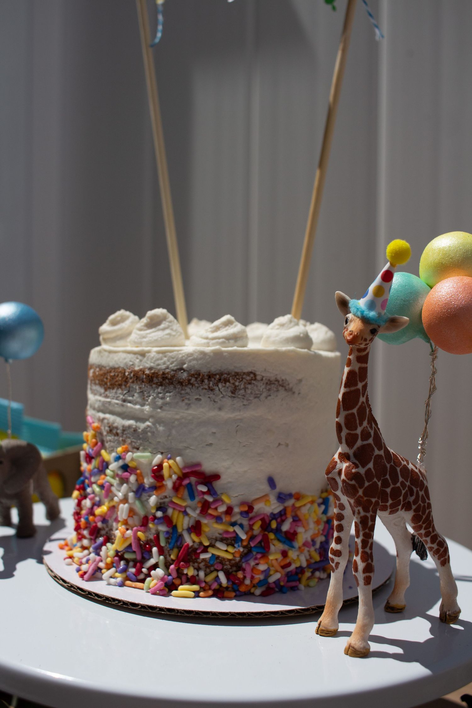 A white frosted birthday cake decorated with colorful sprinkles and a toy giraffe holding pastel balloons on the side.