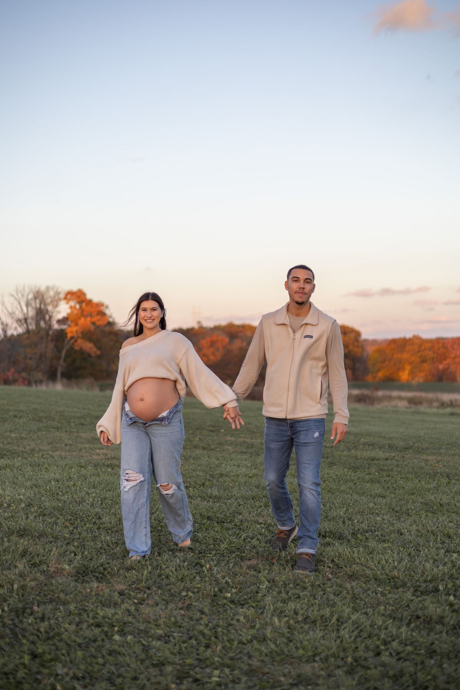 Couple in casual outfits walking playfully in a green field during golden hour sunset.