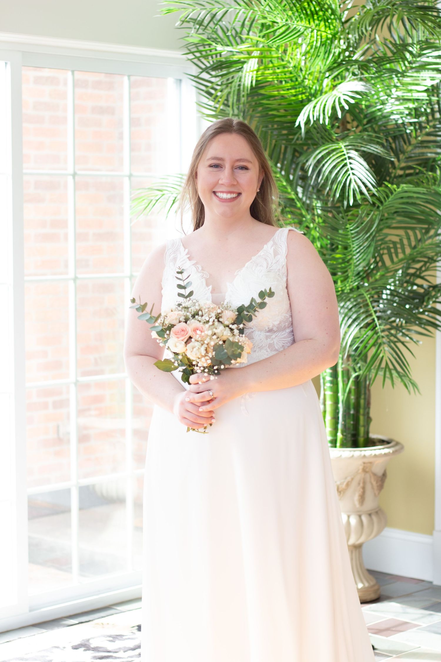 A bride in a flowing white V-neck gown holds a peach and cream floral bouquet while standing near tropical greenery and windows.