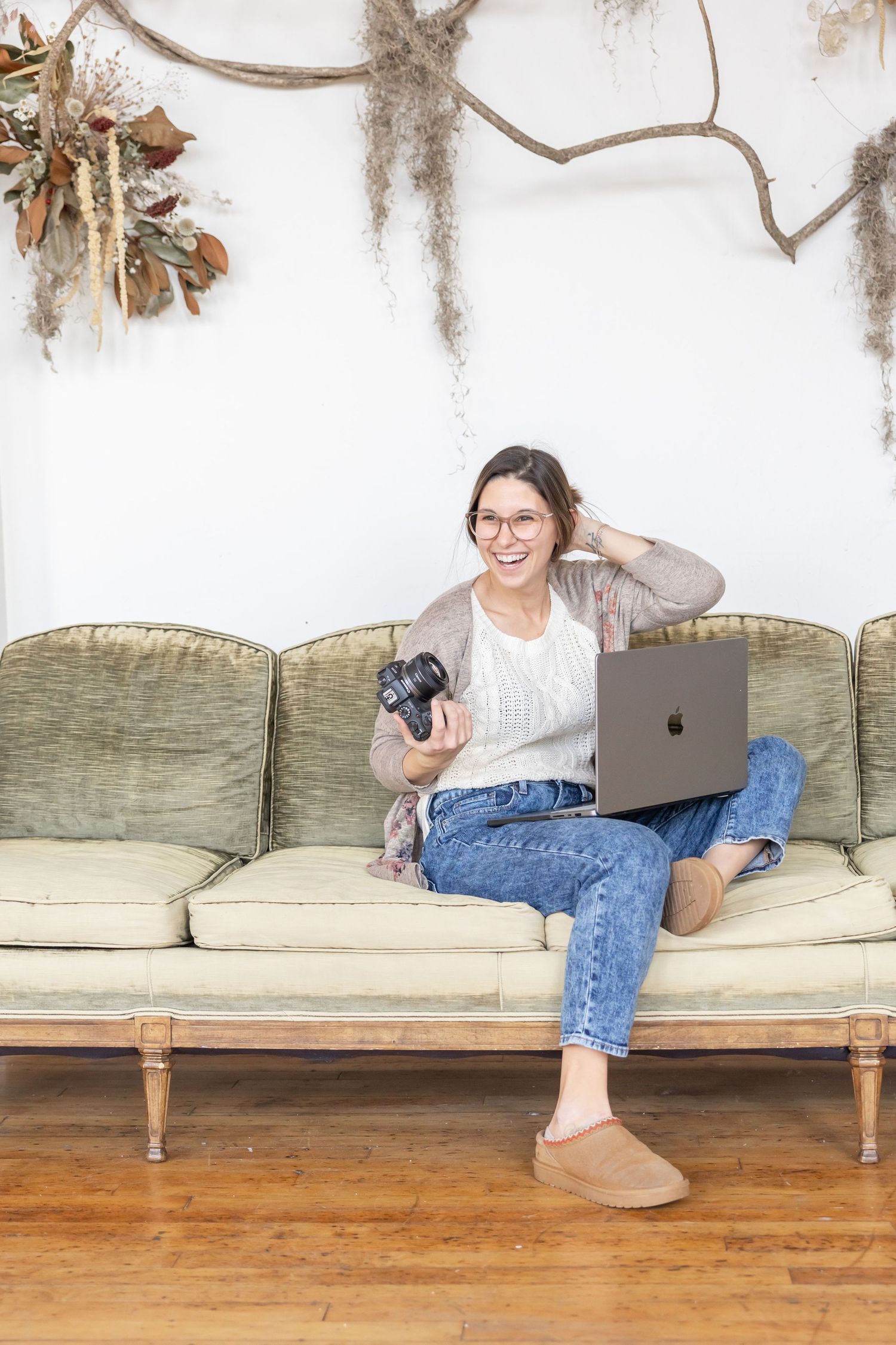 Casual lifestyle scene showing someone relaxing on a vintage couch while working on a laptop and camera in natural light.