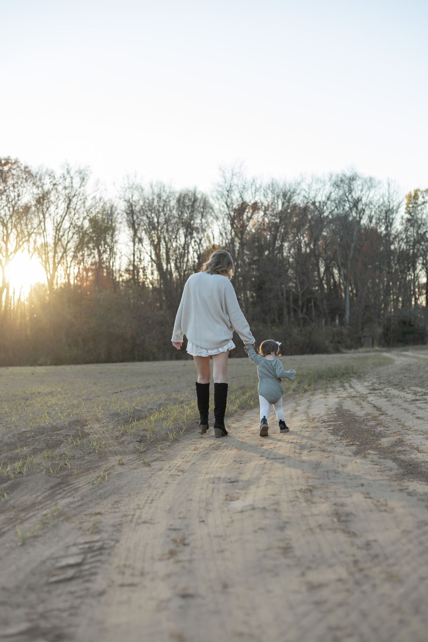 Two figures walk hand in hand down a dirt path at sunset with bare trees in the background.