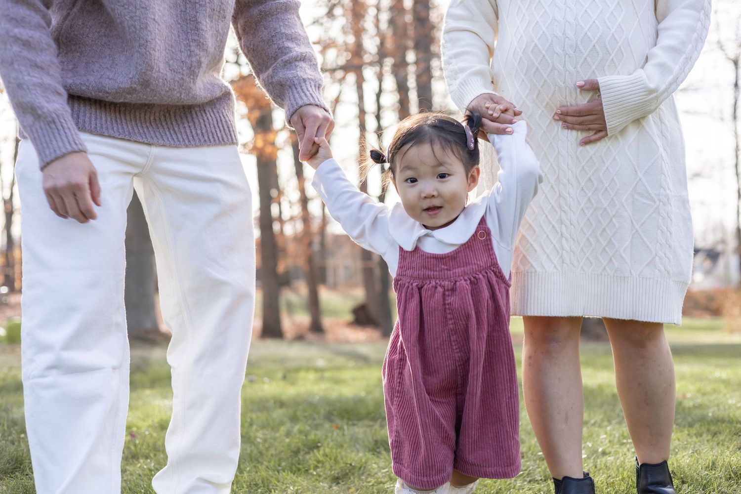 A small child in a burgundy dress holds the hands of their parents while standing on grass during autumn.