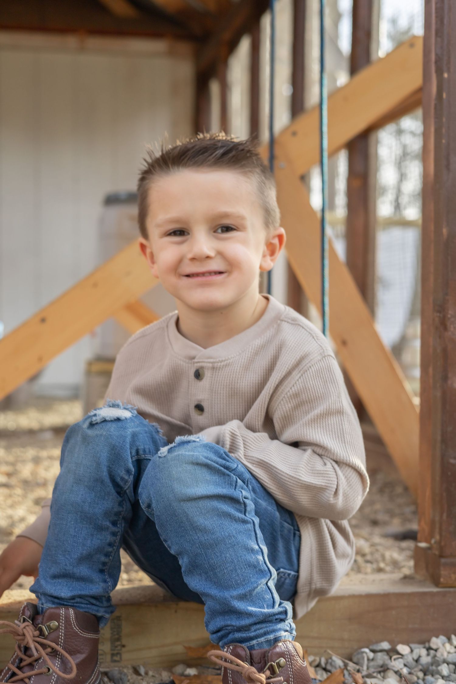 A young child sits on wooden stairs wearing blue jeans, brown boots and a beige henley sweater.