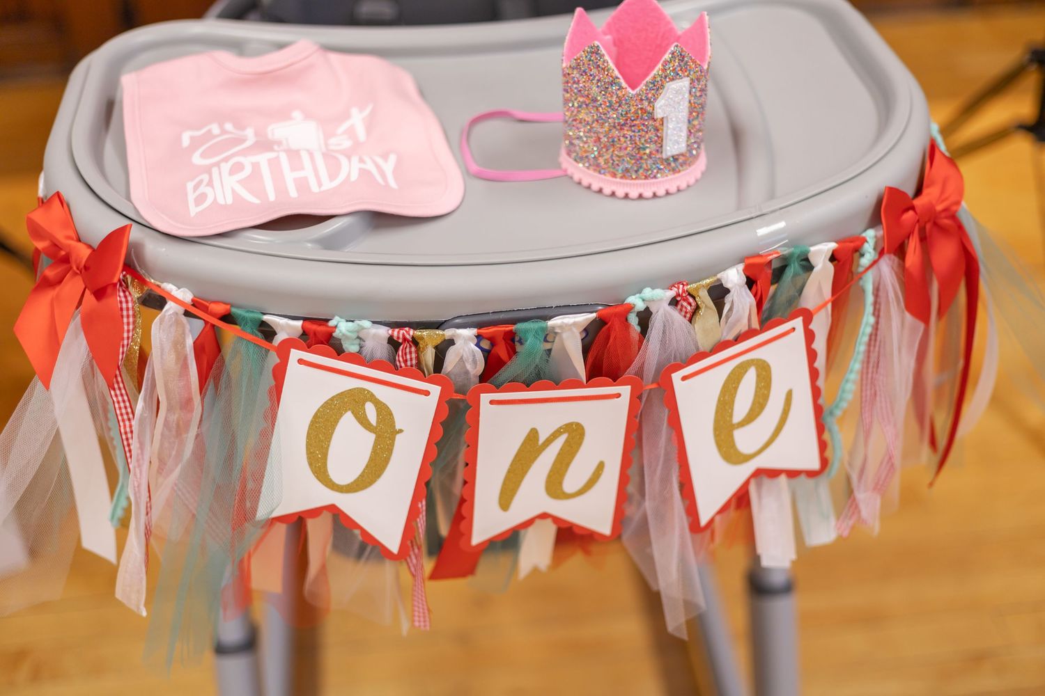 High chair decorated with a festive 'one' banner and pink and gold ribbons for a baby's first birthday celebration.