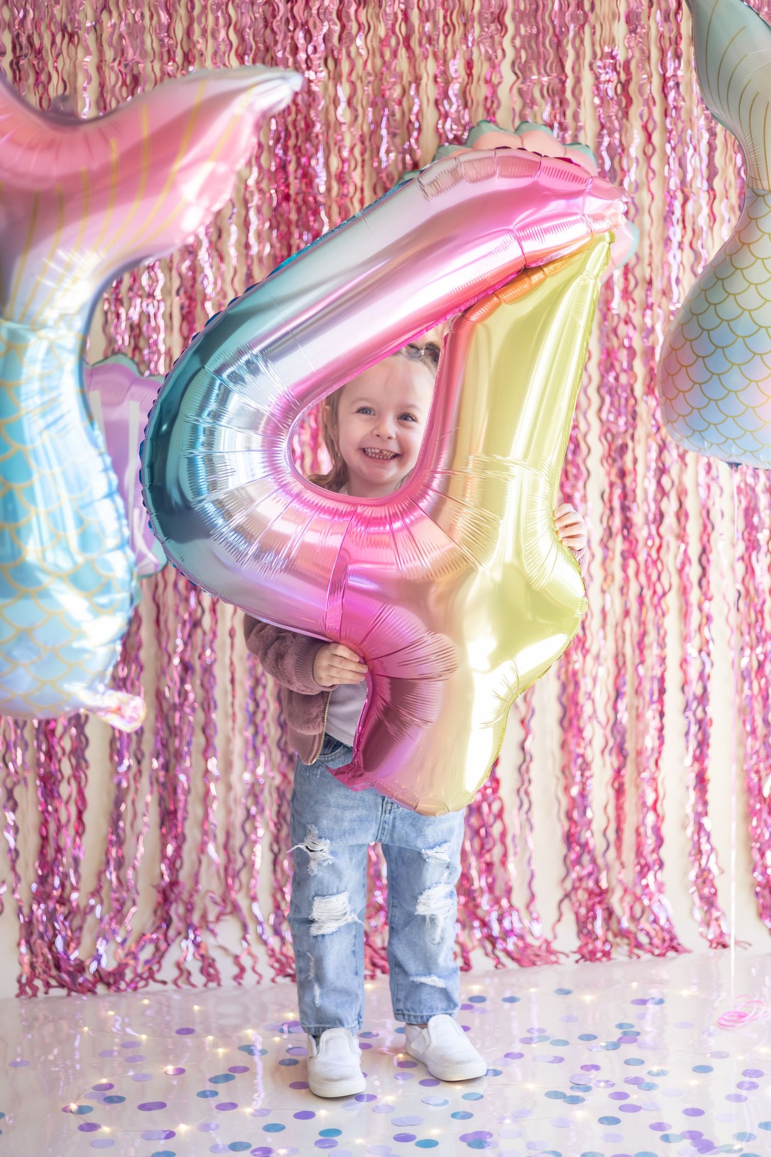 Child stands against pink tinsel backdrop holding number balloons with scattered confetti on white floor.