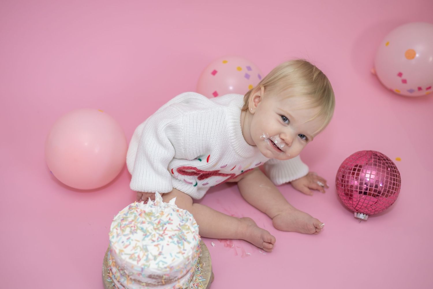 Baby in white dress celebrating first birthday with cake smash photoshoot on pink background with balloons and disco ball.