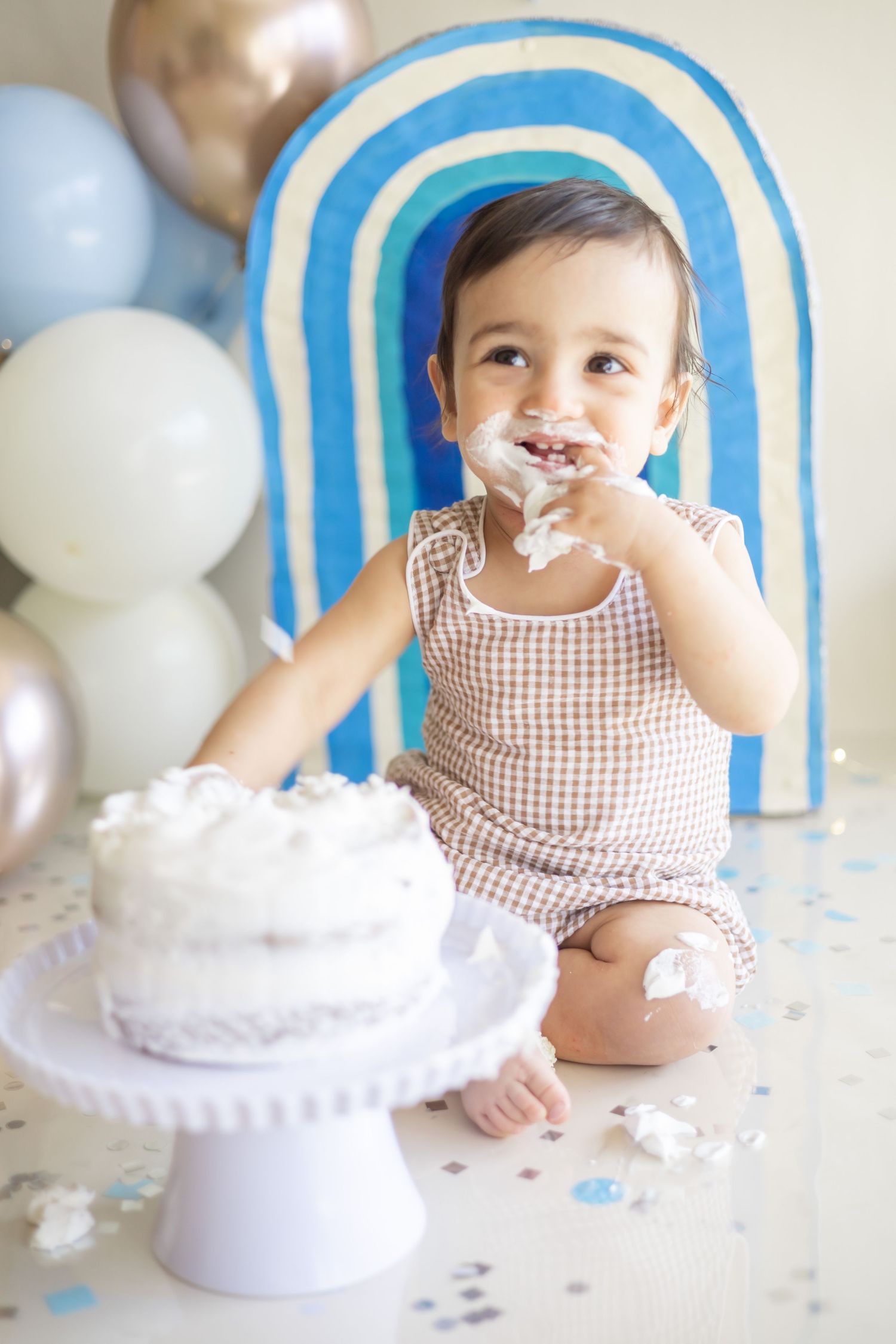 A seated toddler in a brown gingham dress explores and plays with a white cake on a pedestal for their first birthday smash cake.