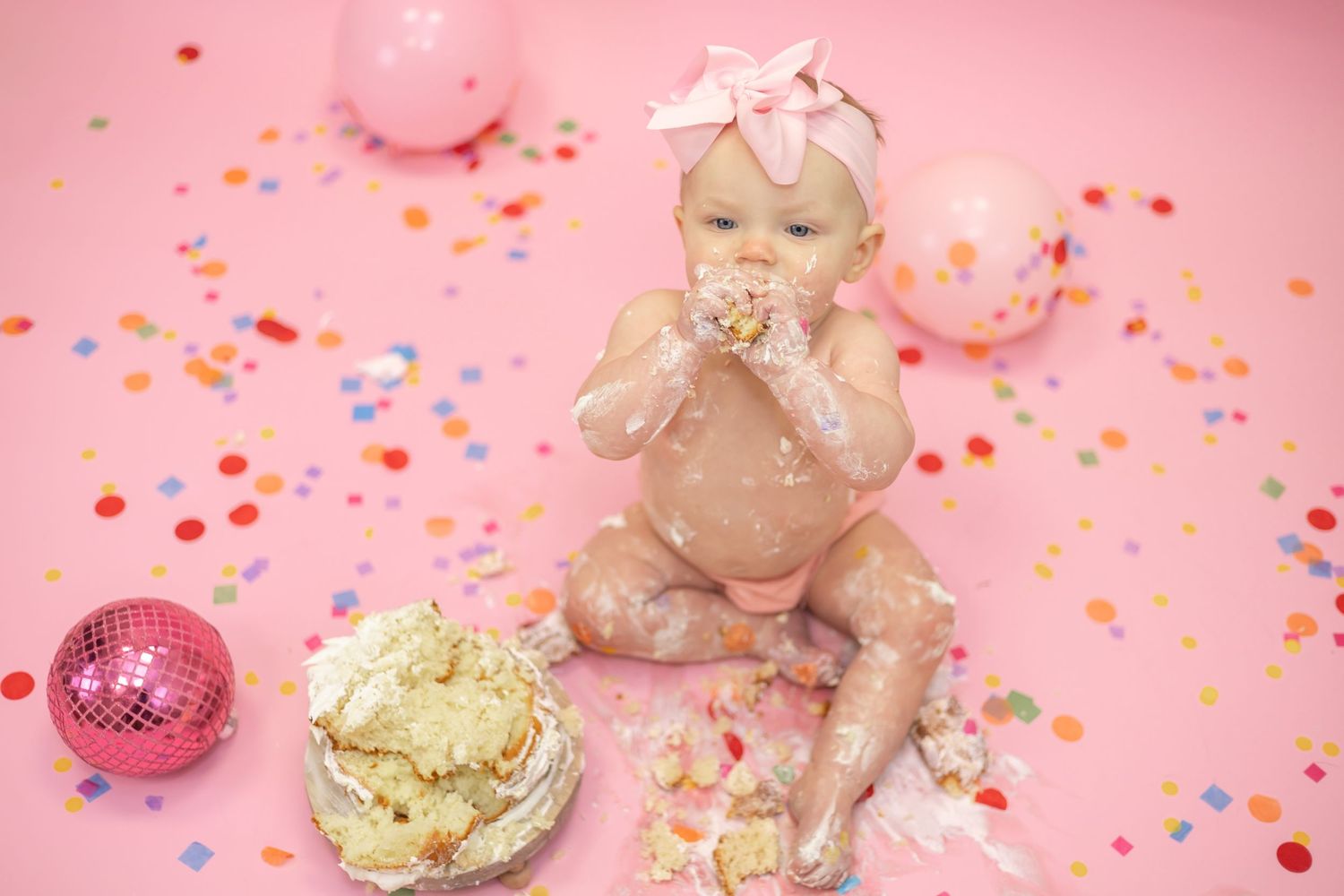 Pink macarons, gold ornaments, disco ball, and plush teddy bear arranged on soft pink background with scattered confetti.
