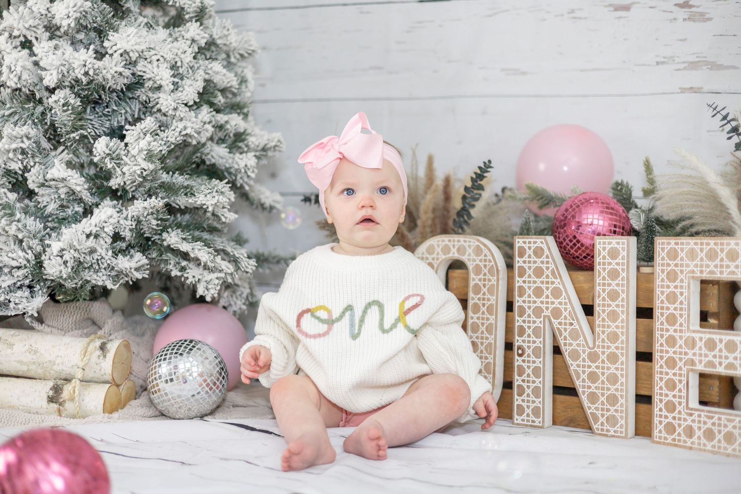 Baby in white onesie sits on rustic wooden floor with winter holiday decor of pink and silver ornaments and sparkly letters.
