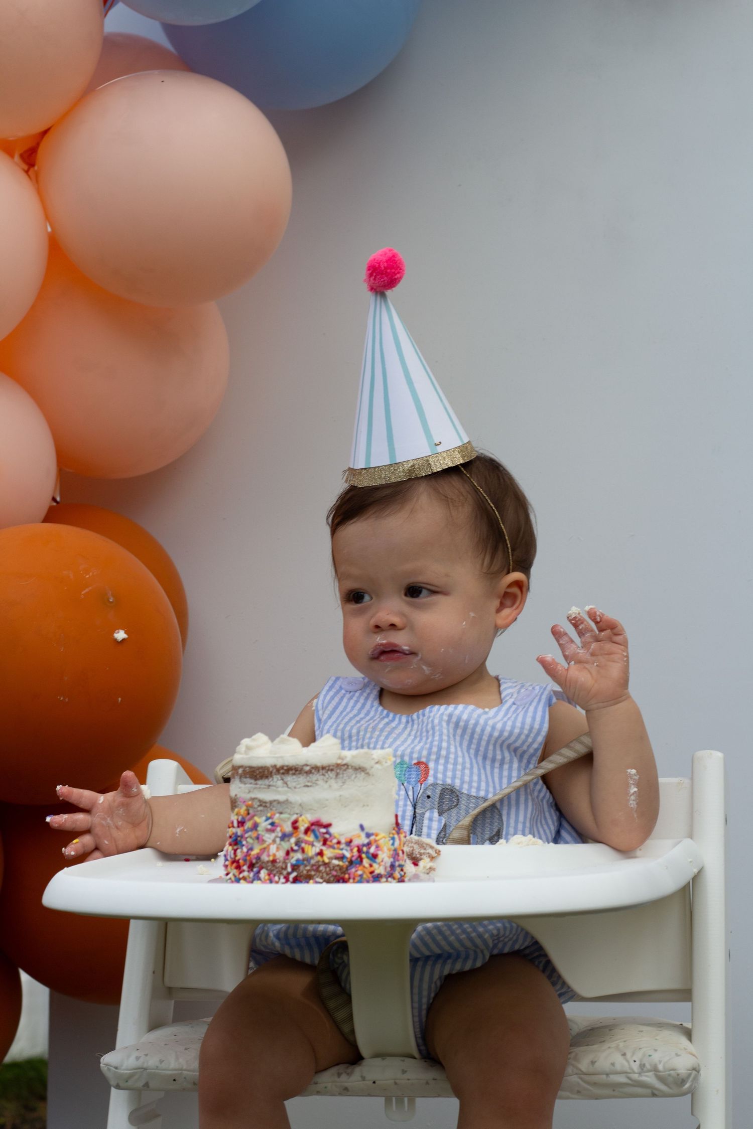 Small child in high chair celebrates birthday with cake and orange and blue balloon decorations.
