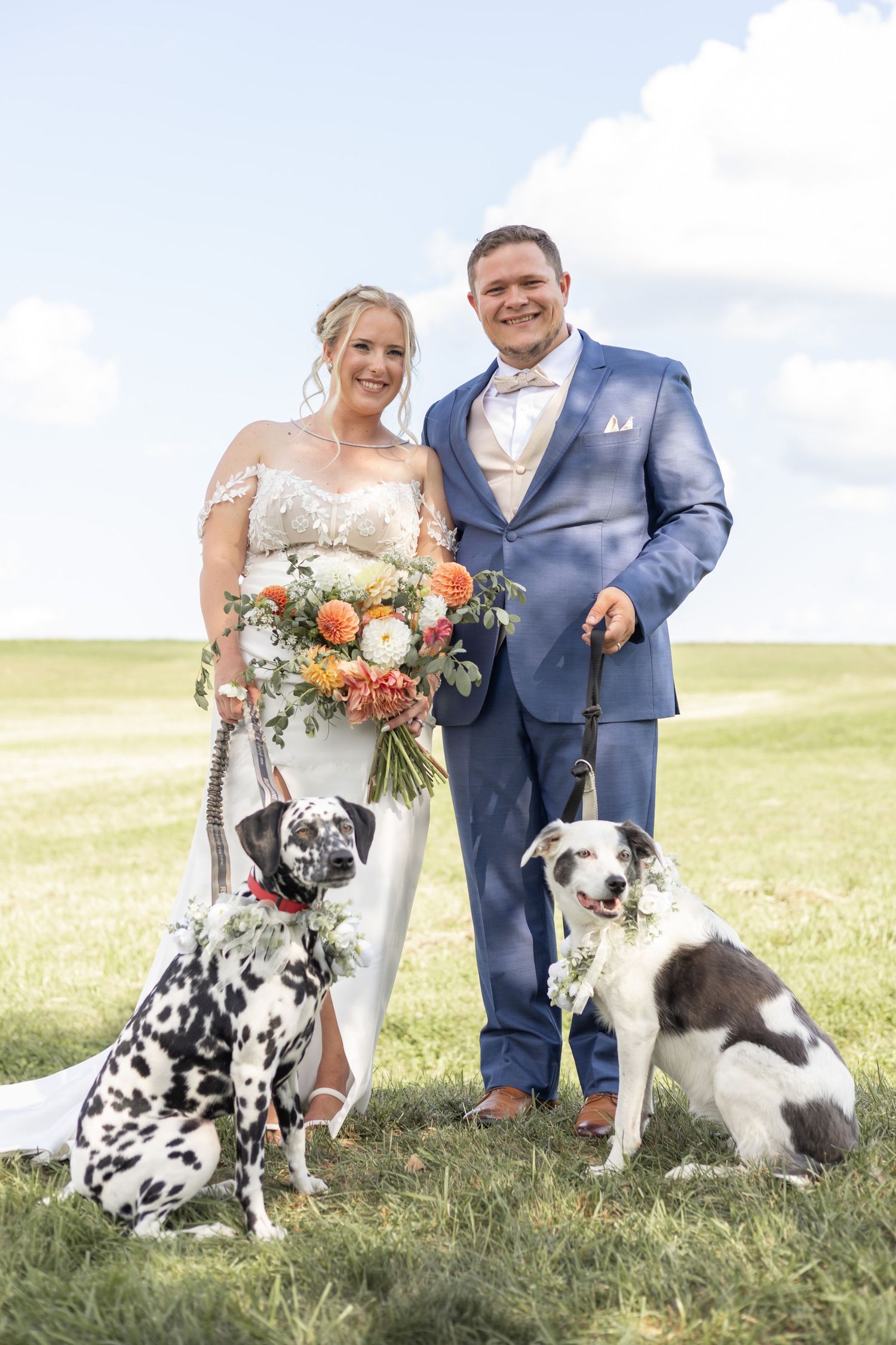Wedding couple posing on grassy hillside with their spotted Dalmatian dog wearing floral collar in outdoor setting.