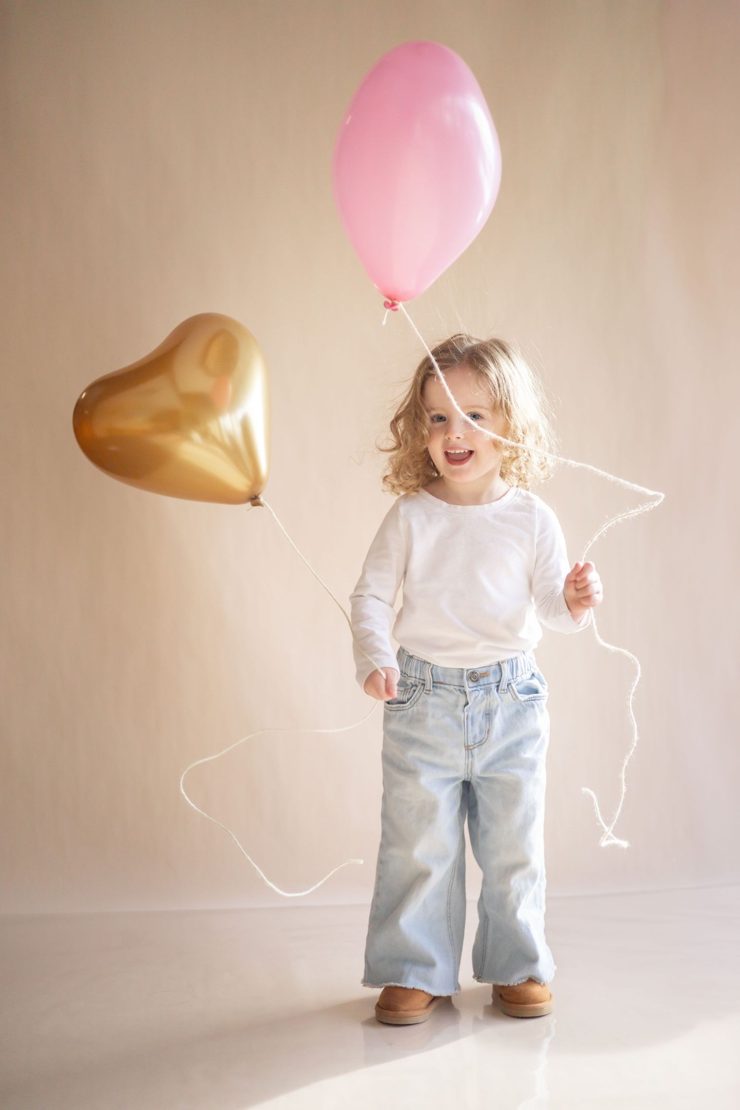 A child wearing blue jeans and white sweater holds pink and gold heart-shaped balloons against a neutral background.