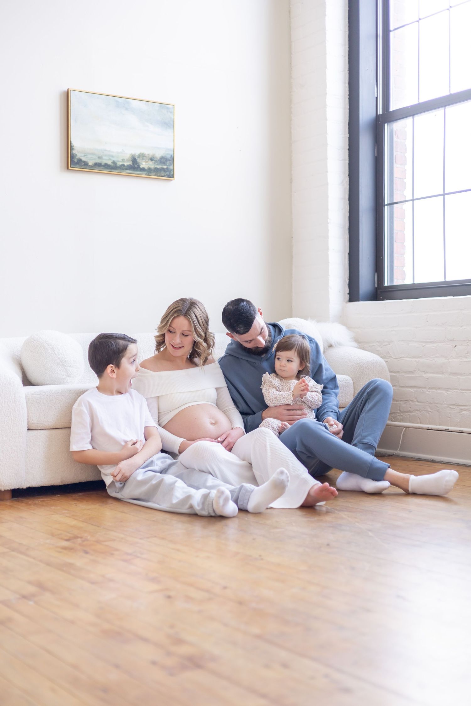 A family sits together on a hardwood floor in a bright minimal room with white walls and a framed landscape painting.