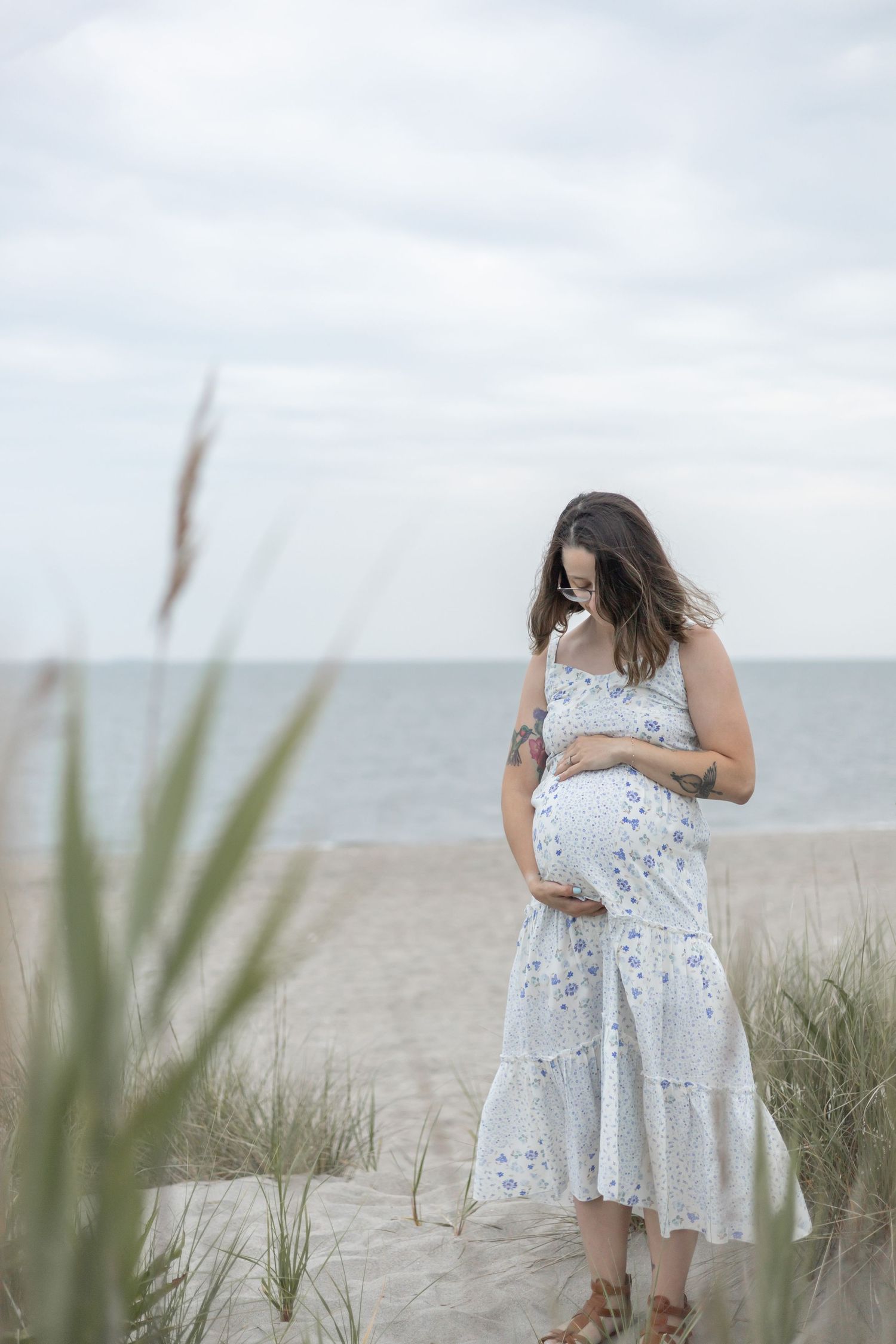 Expectant mother in a flowy white floral dress stands thoughtfully among beach grass with the ocean in the background.