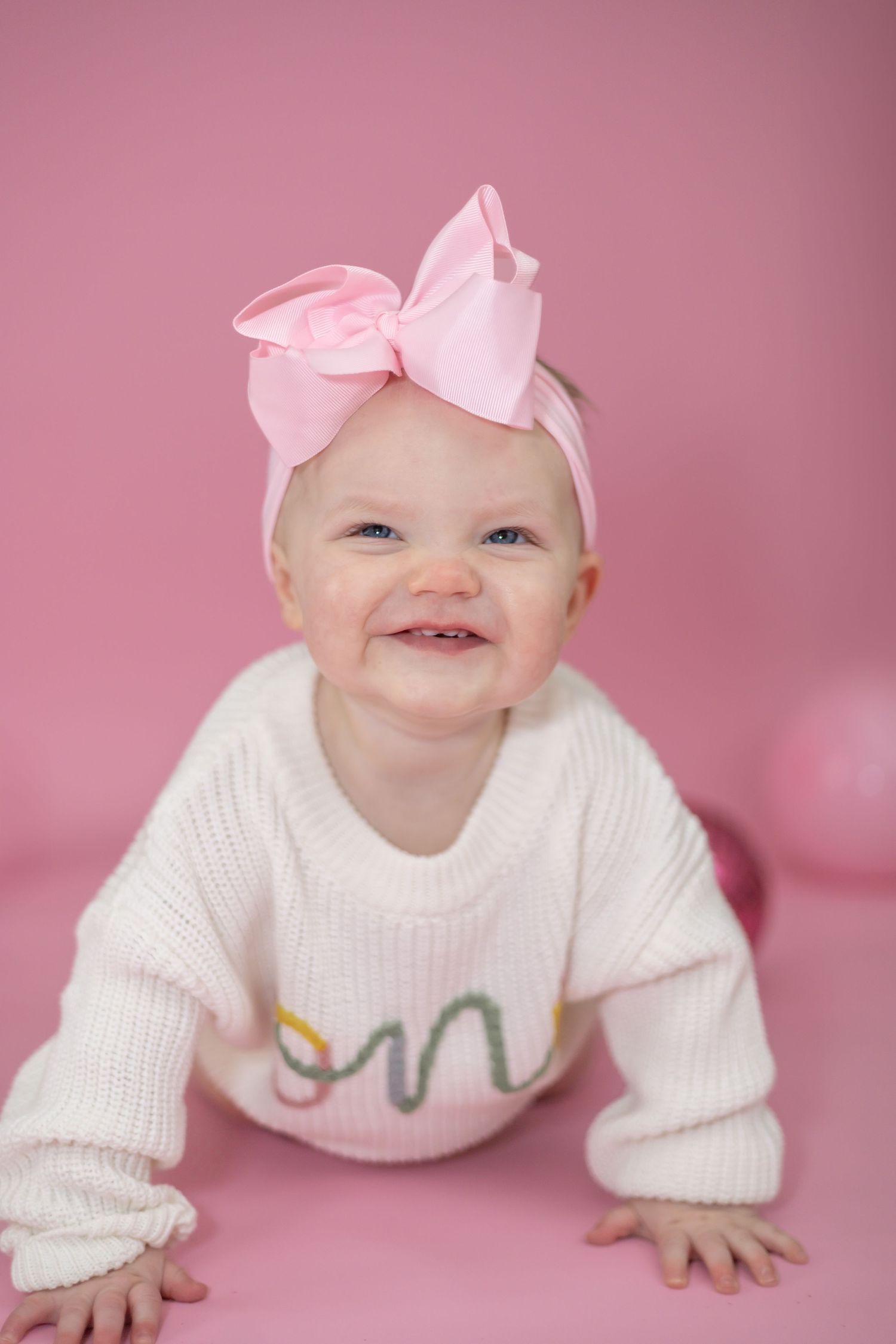 Baby wearing white sweater and pink bow headband smiling against pink background.