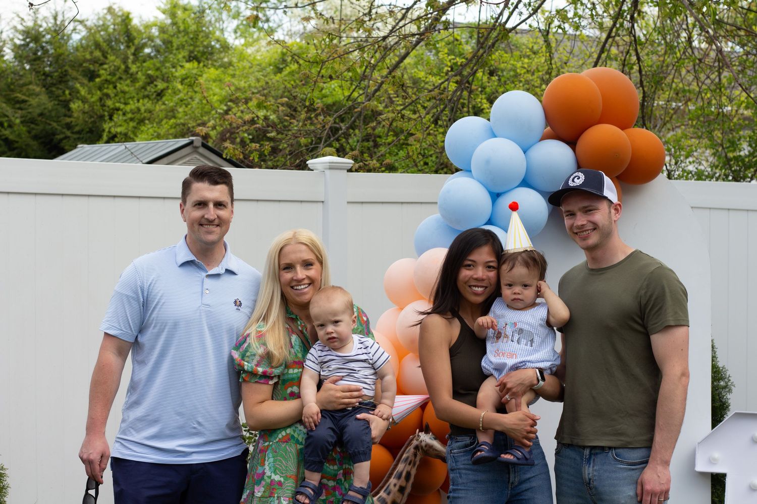 Group gathered outdoors with blue and orange balloons against white fence for celebration event.