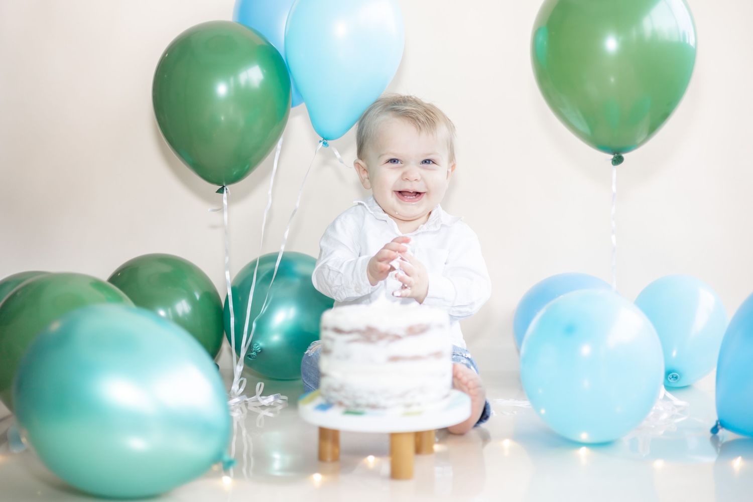 First birthday celebration setup with blue and green balloons surrounding a small white cake on the floor.