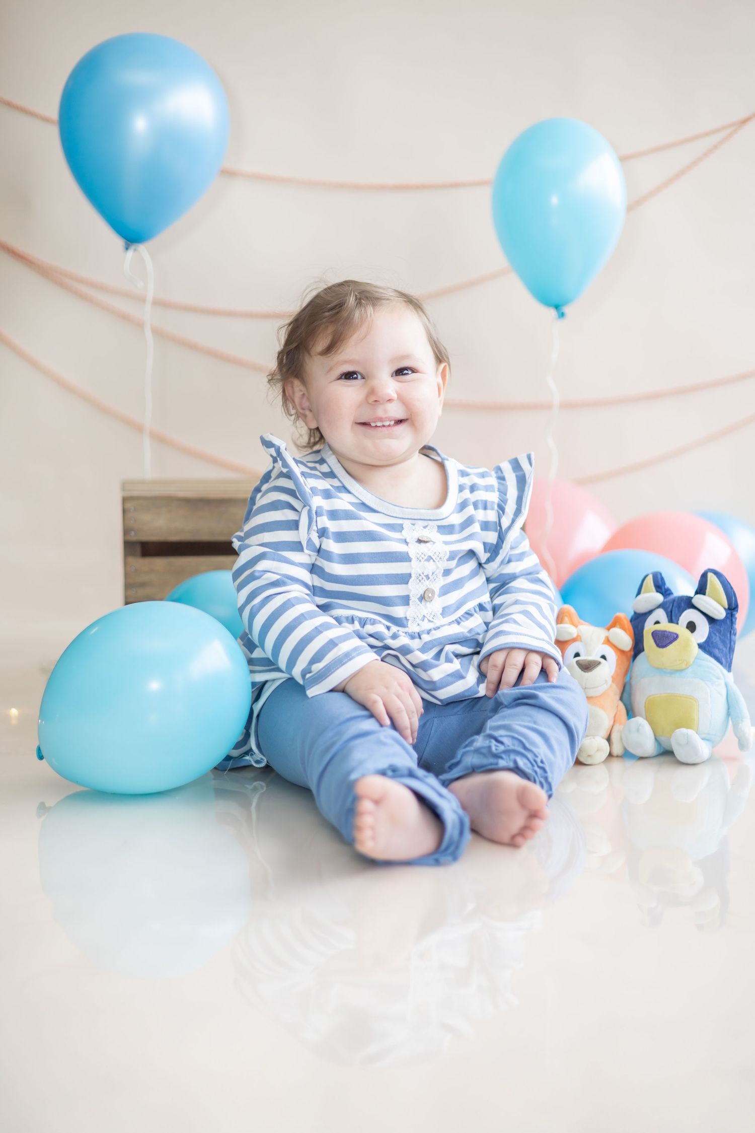 Adorable baby in striped outfit sitting with blue balloons and plush toys against white background.