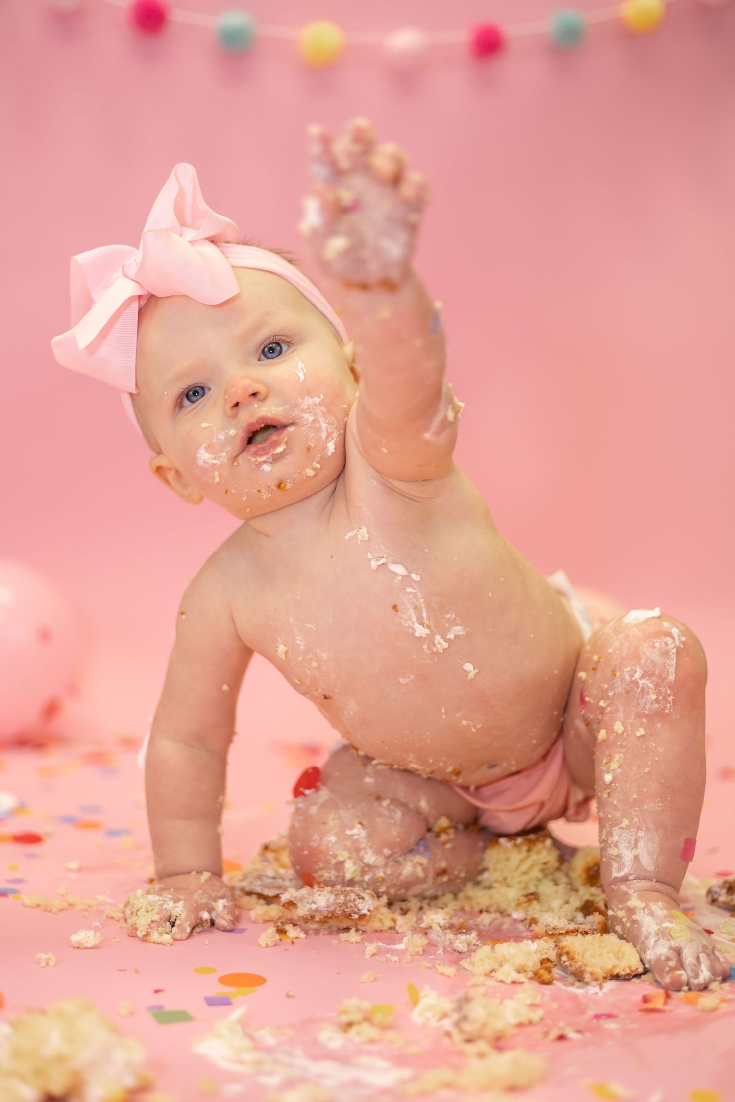 Baby with pink bow having fun during cake smash photo session with colorful party decorations.