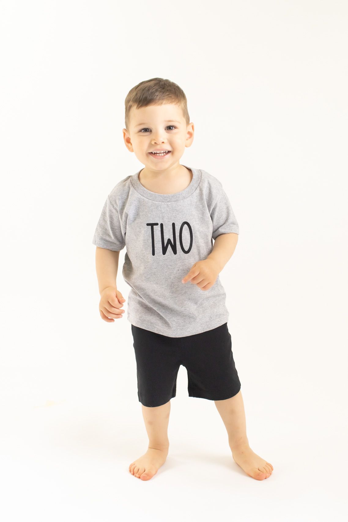 Toddler wearing gray t-shirt with 'TWO' text and black shorts plays on white background.