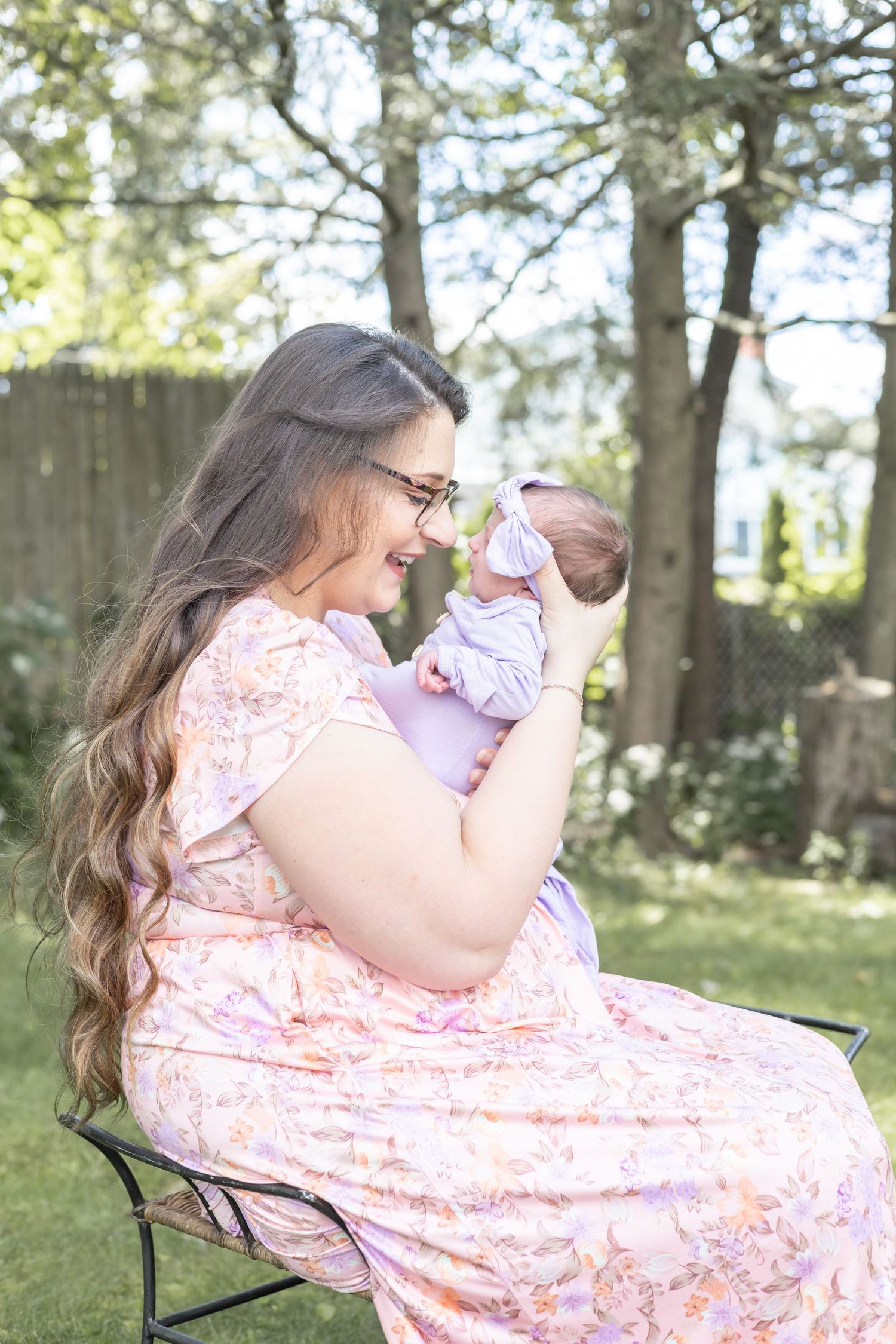 A mother in a floral pink dress holds her tiny baby while sitting in a garden chair on a sunny afternoon.