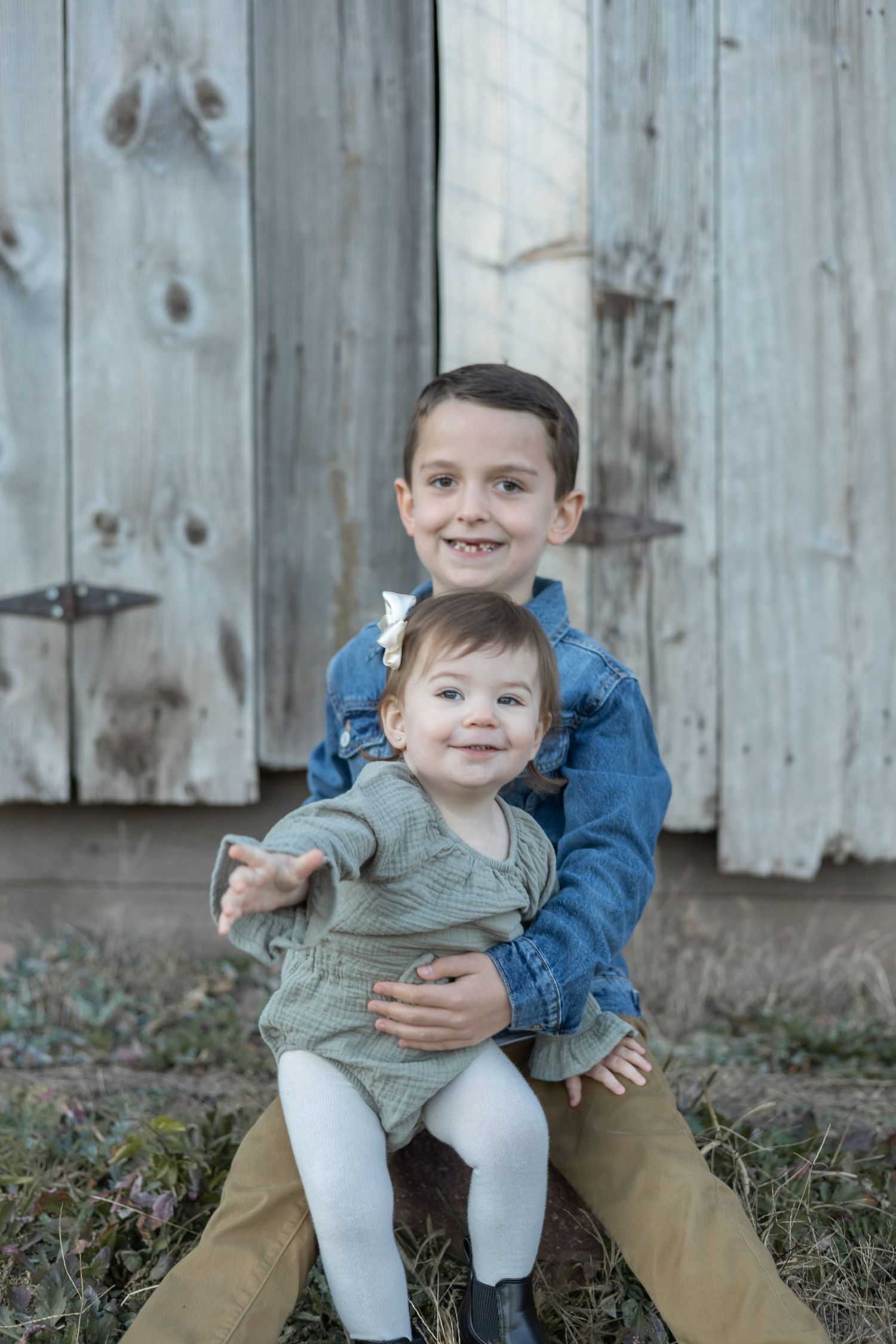 Two children in denim and khaki sitting together against a weathered wooden door.