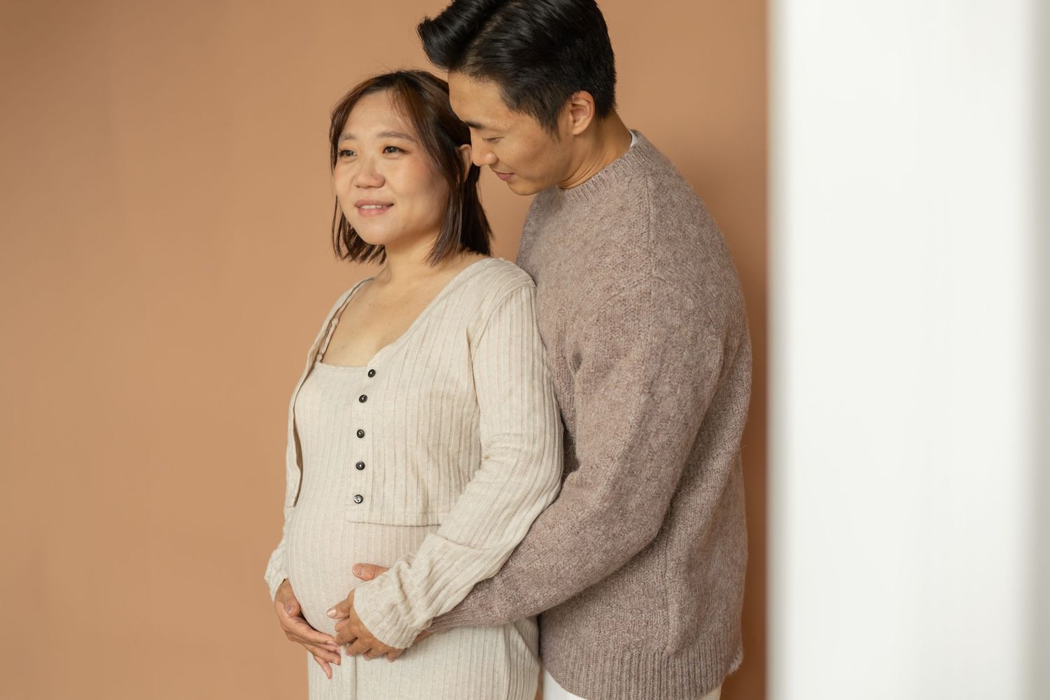 Expectant couple sharing tender moment against beige backdrop as they gently cradle a pregnant belly.