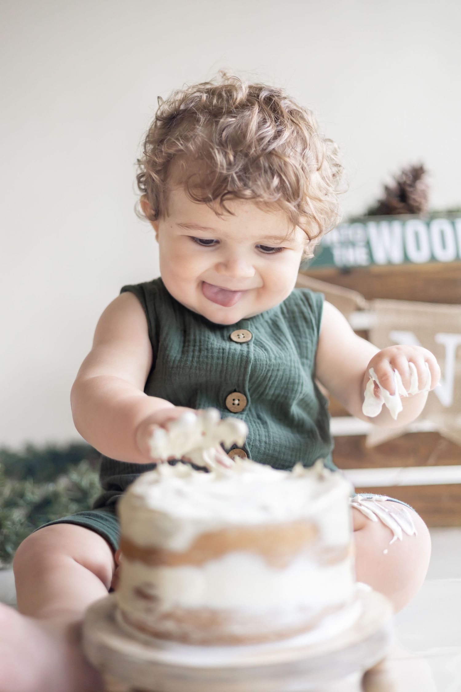 A toddler in a green sleeveless outfit reaches for a white frosted naked cake while smiling with joy.