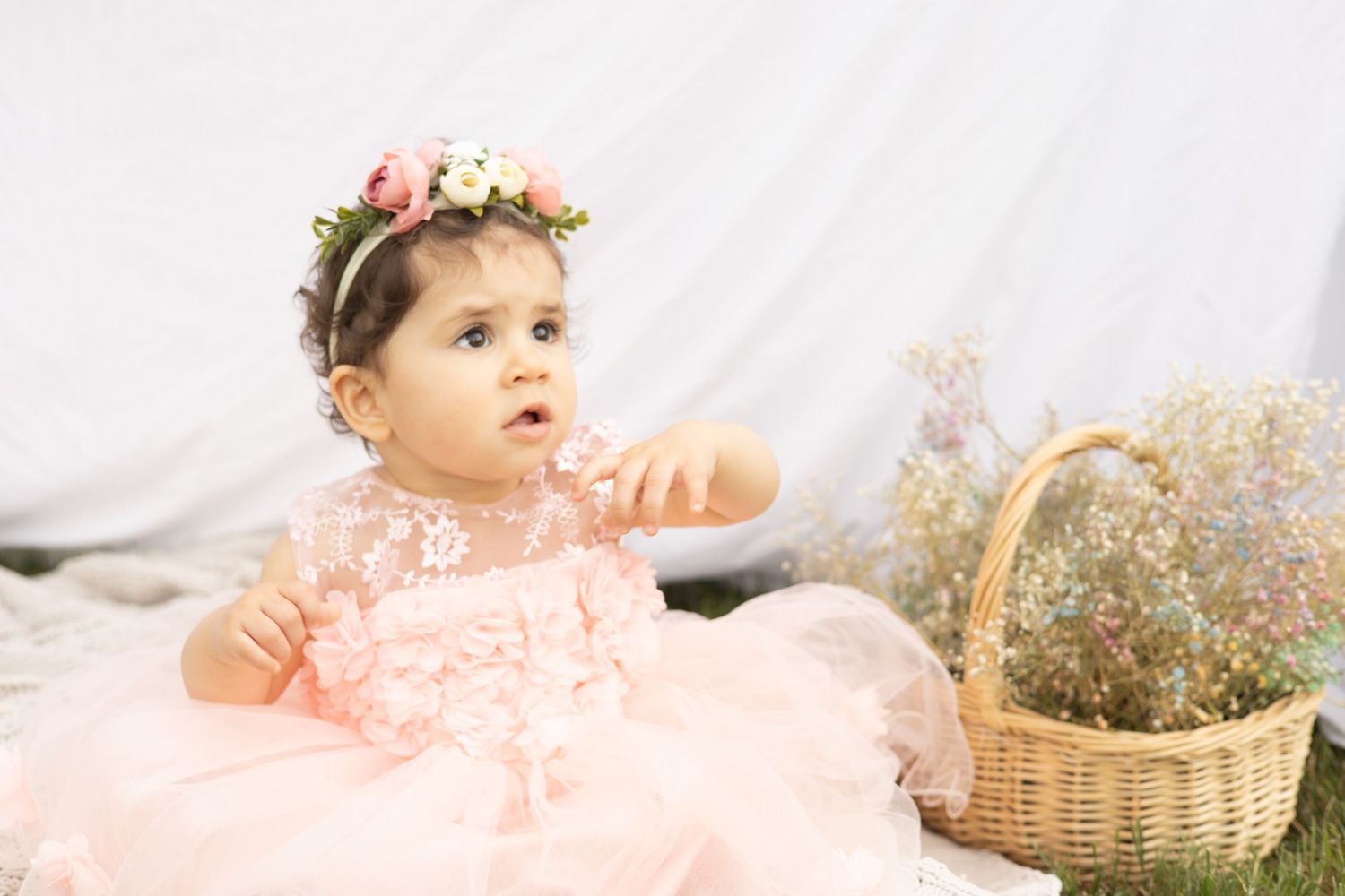 A small figure in a pink tulle dress and floral crown sits beside a wicker basket with dried flowers.