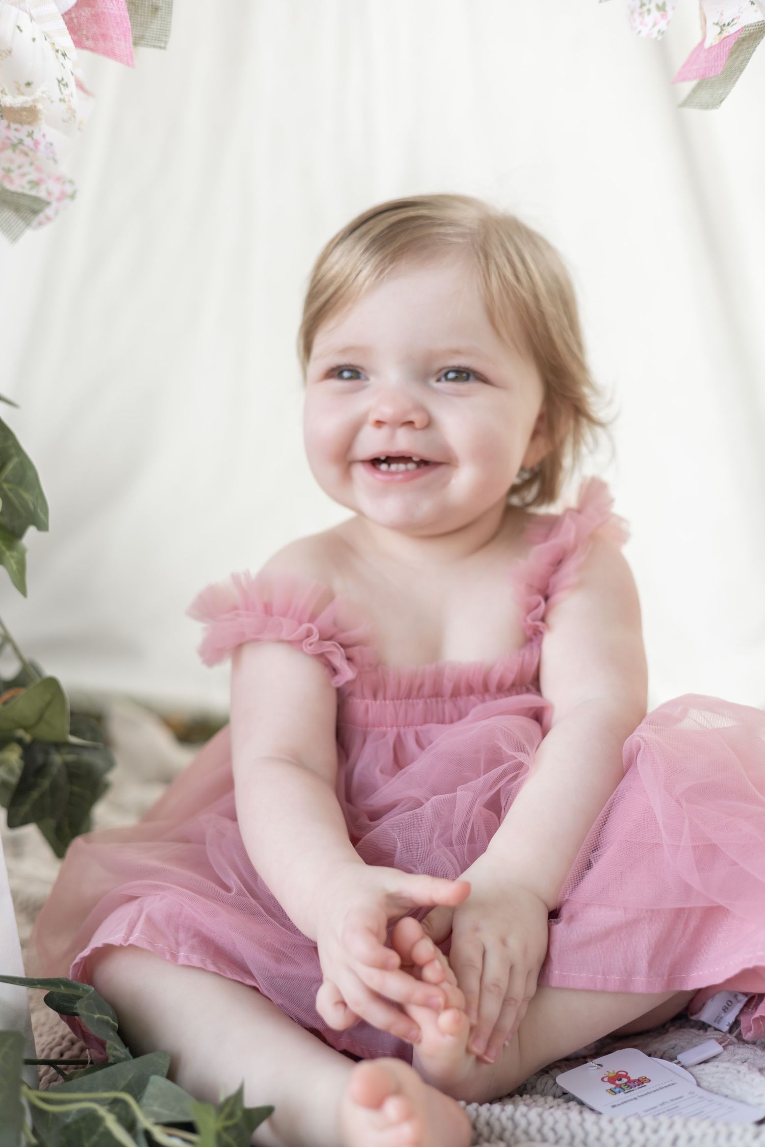 A happy toddler wearing a pink tulle dress sits on a white backdrop surrounded by floral decorations.