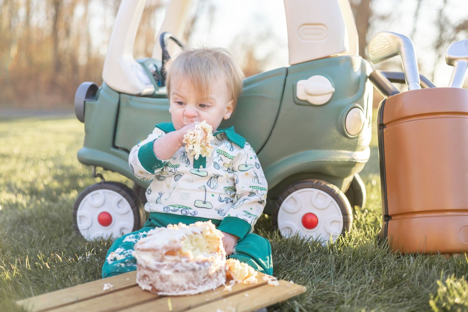 A one-year-old baby sits in the grass wearing a colorful birthday bib while messily eating their first birthday cake.