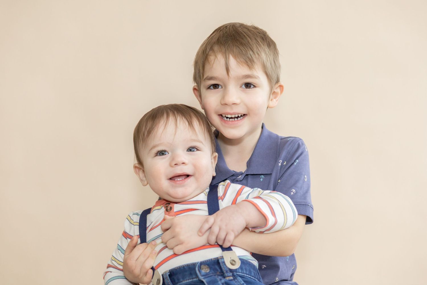 Two young children embrace while smiling against a beige backdrop.