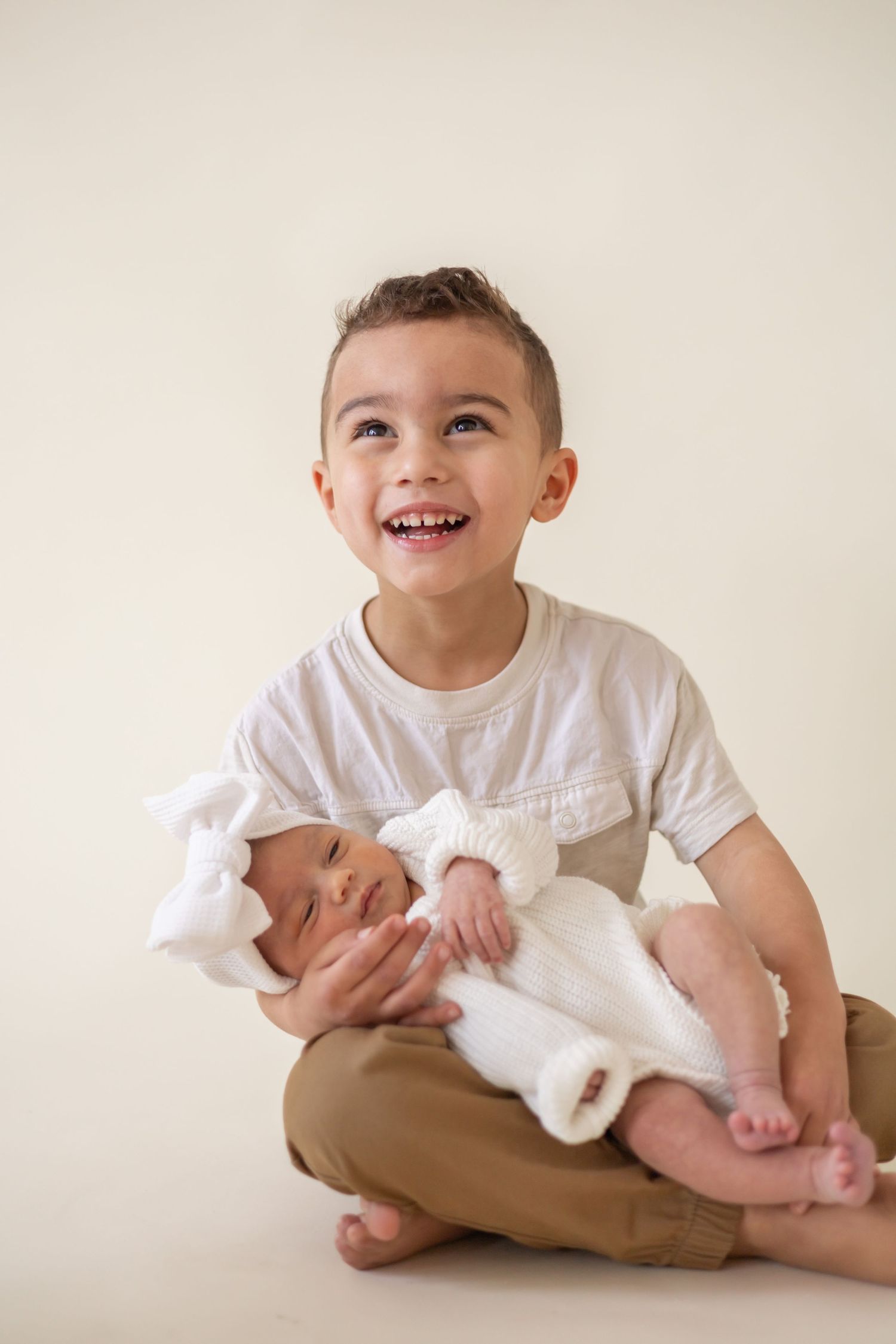 A young child sits cross-legged while holding and smiling at a newborn baby wrapped in white.