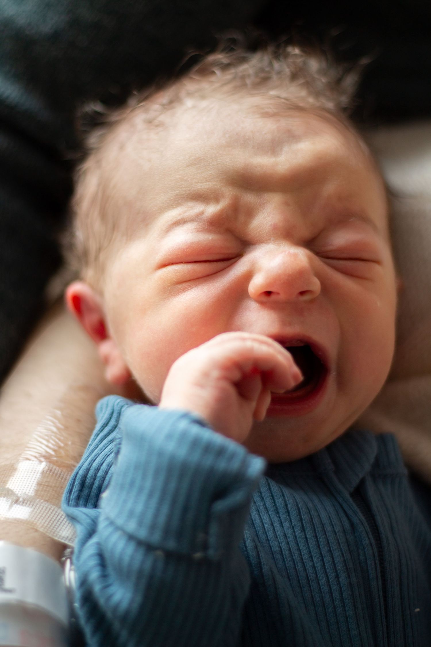 A newborn baby in a blue sweater is yawning while resting on a cozy surface.