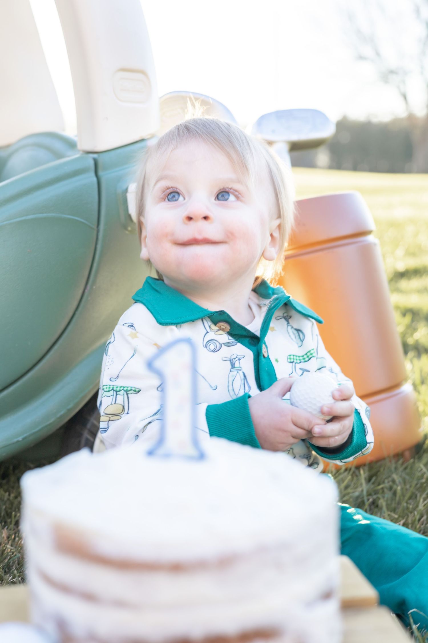 A toddler in a white and green outfit smiles while celebrating their first birthday with a cake outdoors.